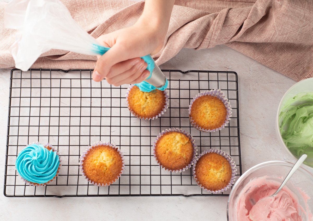 A person is decorating gluten-free vanilla cupcakes with blue buttercream icing on a wire cooling rack, with additional pastel frosting in bowls nearby, suggesting preparation for Easter-themed treats.
