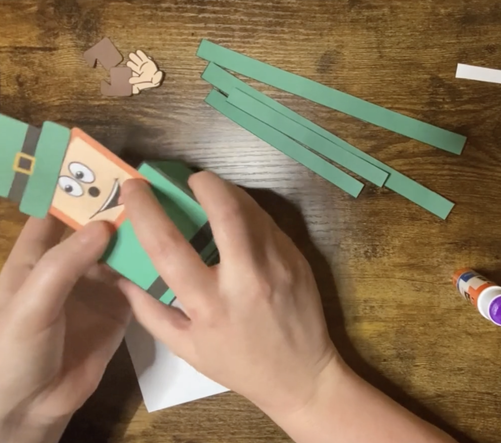 A person is applying glue to a paper while assembling a three-dimensional leprechaun craft, suitable for St Patrick's Day activities for preschool and kindergarten.
