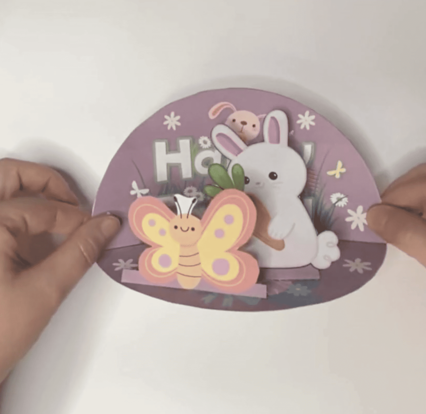 A pair of hands holds a DIY Easter card featuring a bunny and a butterfly, surrounded by flowers, designed for kids as part of spring crafts and activities.
