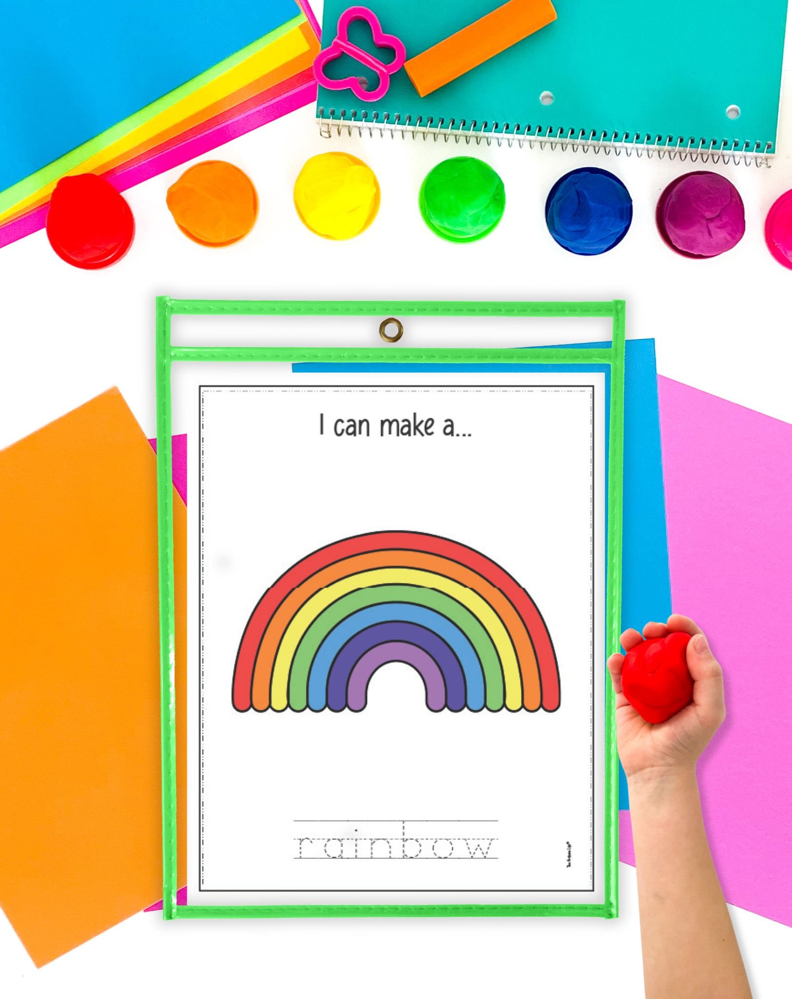 A child's hand holding a red playdough ball is positioned next to a colorful activity mat with a rainbow design and various play dough containers, surrounded by bright sheets of paper, showcasing materials for spring preschool playdough activities.
