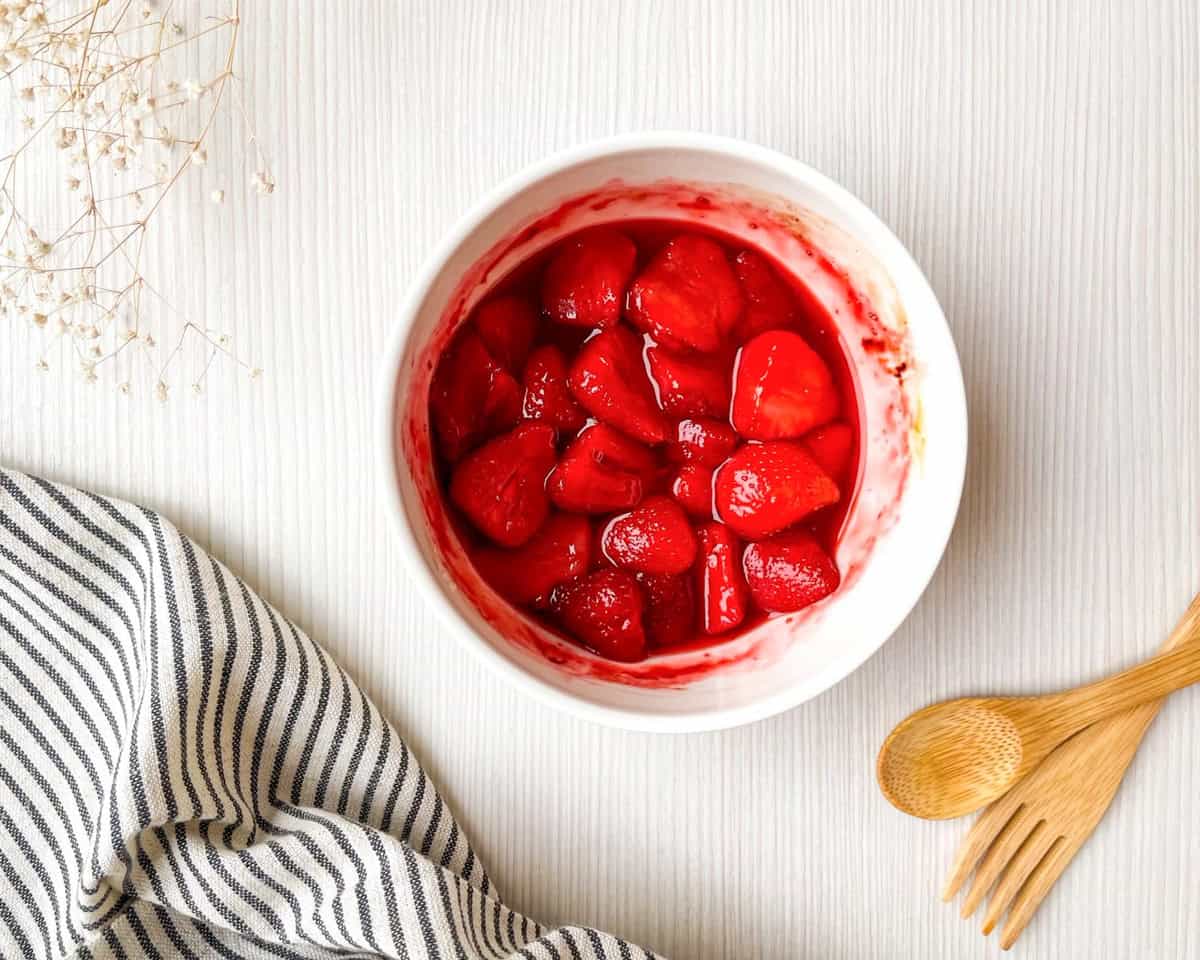 A bowl of roasted strawberries is placed on a light background, accompanied by wooden utensils and a striped cloth, suggesting a preparation for a breakfast or brunch idea, such as a strawberry Dutch baby pancake recipe.
