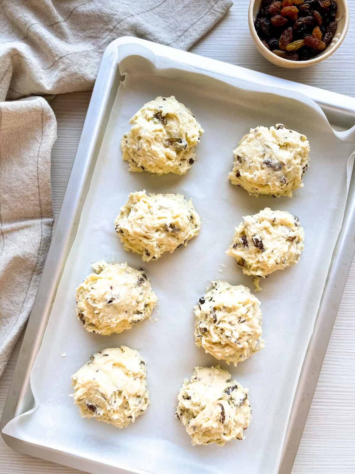 A tray lined with parchment paper holds eight uncooked buttermilk scones mixed with raisins, alongside a small bowl of additional raisins and a cloth napkin.
