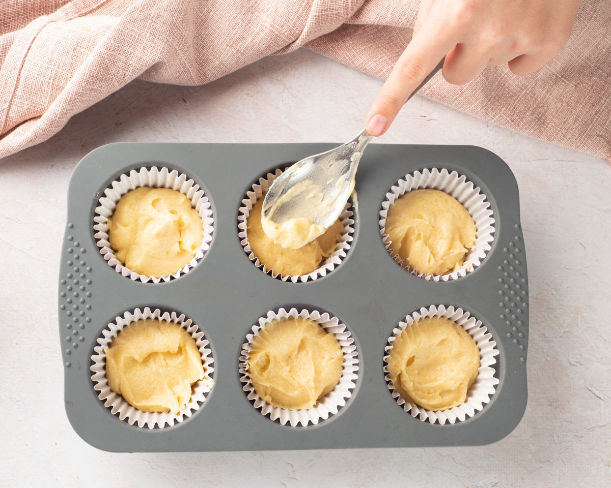 A hand is placing a paper cupcake liner into a gray muffin tray, surrounded by ingredients such as flour and an egg, suggesting preparation for making gluten-free cupcakes, possibly for an Easter celebration.
