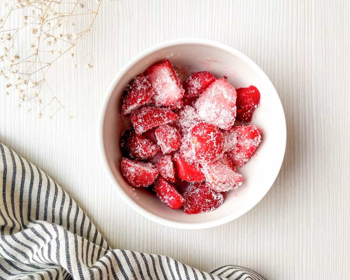 A white bowl filled with sugared strawberries is placed on a light surface next to a striped cloth, suggesting ingredients for a strawberry Dutch baby pancake recipe or breakfast brunch ideas.
