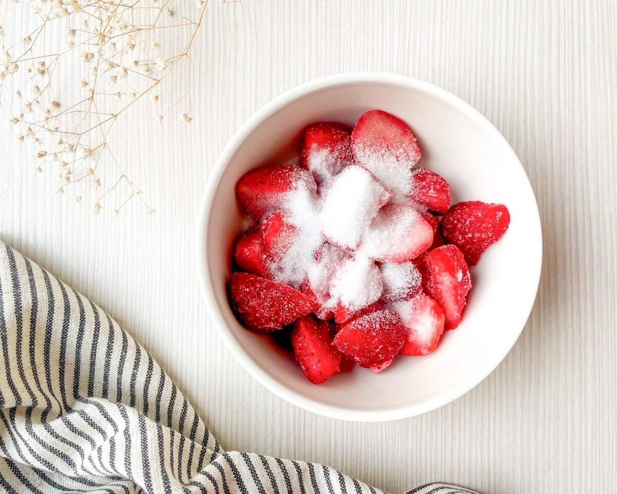 A bowl of sliced strawberries sprinkled with sugar sits on a light wooden surface, accompanied by a striped cloth, suggesting a preparation context for a strawberry Dutch baby pancake recipe.
