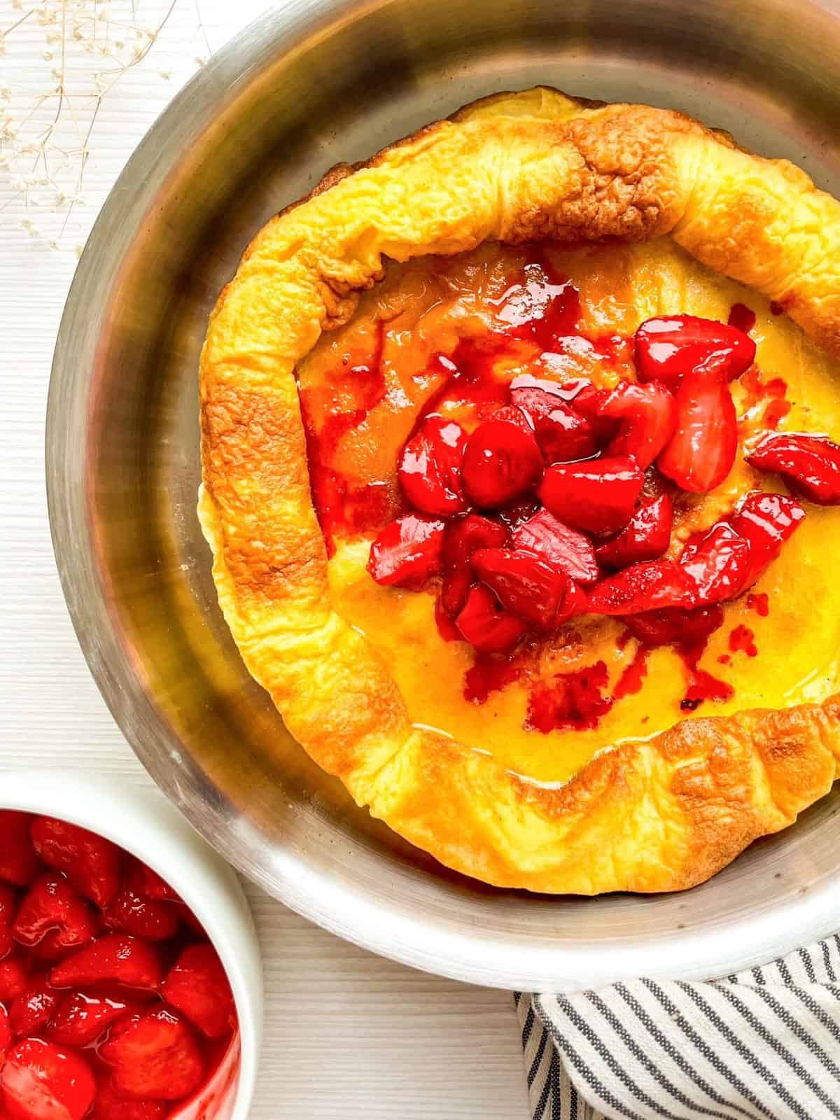 A Dutch baby pancake topped with strawberry sauce is displayed in a metal pan, accompanied by a bowl of additional strawberries, highlighting a breakfast or brunch idea.
