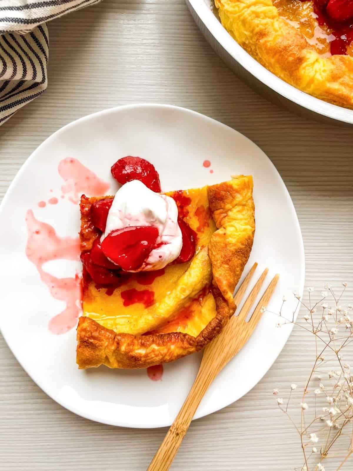 A slice of strawberry Dutch baby pancake is served on a white plate, topped with whipped cream and strawberry sauce, alongside a striped cloth.
