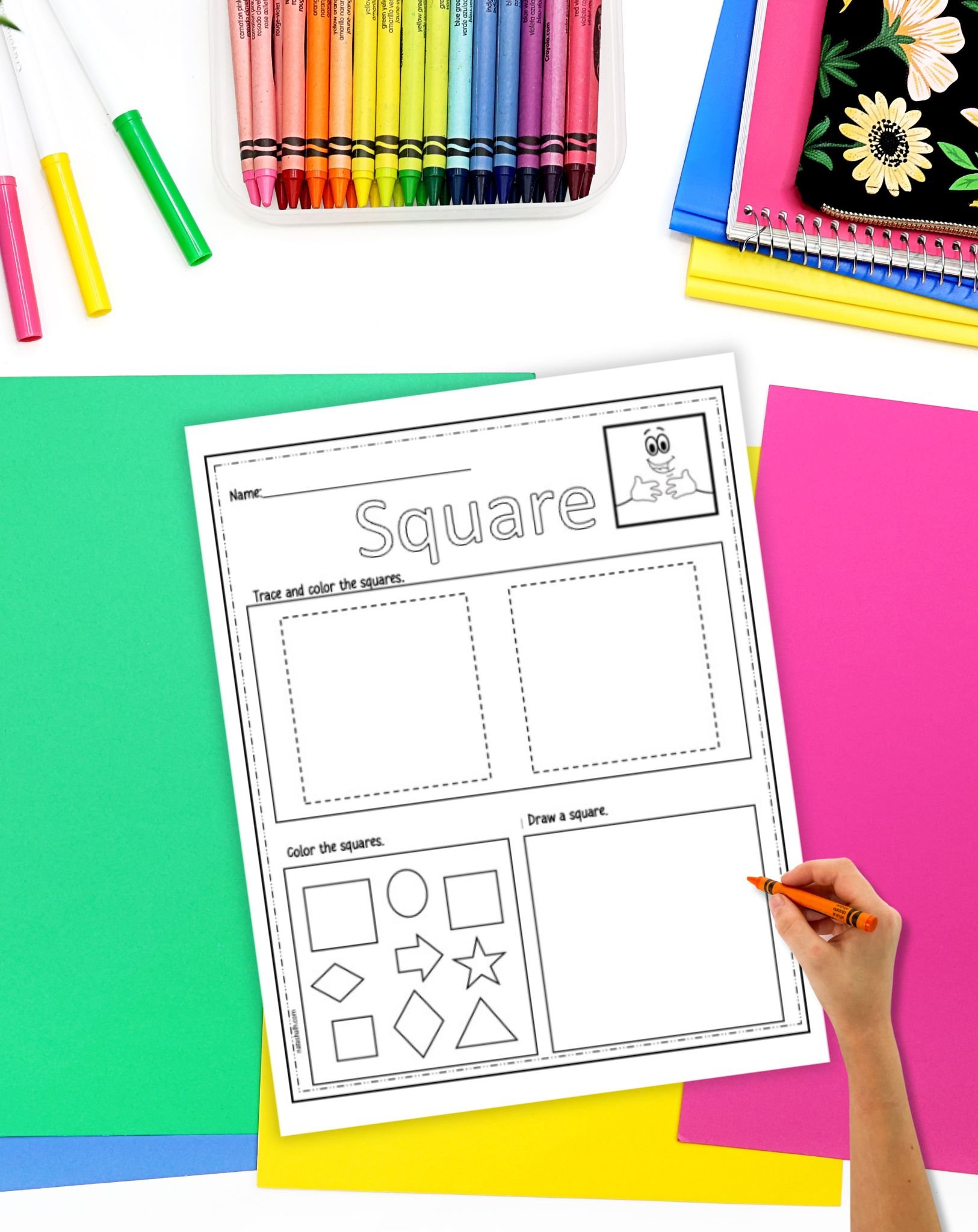 A hand is using an orange crayon to trace shapes on a square tracing worksheet designed for preschool activities, surrounded by various colored papers and a container of crayons.
