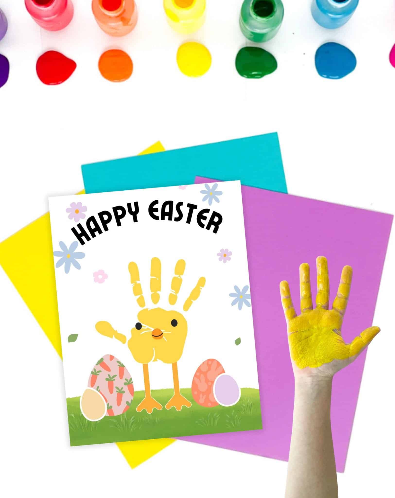 A child's hand with yellow paint is raised next to a festive "Happy Easter" card featuring a handprint chick, surrounded by colorful craft supplies and paper, illustrating an Easter handprint craft activity for toddlers.
