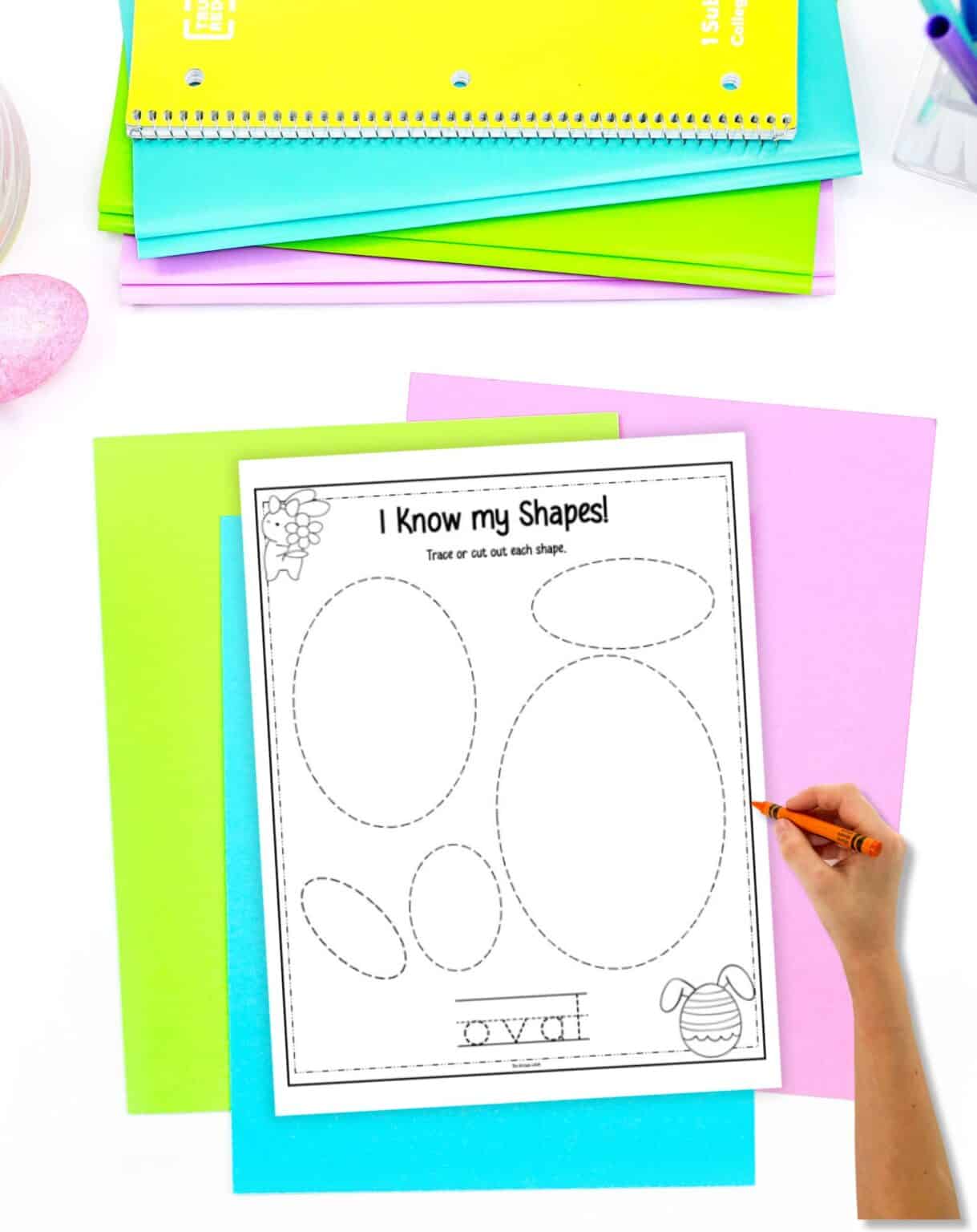 A hand holds a crayon over a worksheet titled "I Know my Shapes!" featuring star shapes for tracing, surrounded by colorful papers and crayons, suitable for Easter learning activities for preschool.
