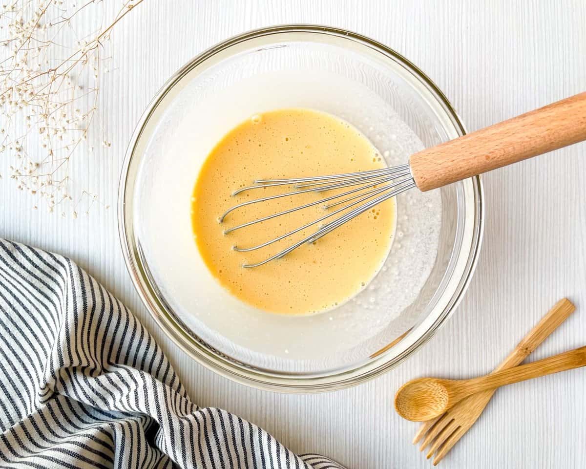 A glass bowl containing a whisked mixture of eggs and milk is shown, alongside striped fabric and wooden utensils, suggesting preparation for a Dutch baby pancake recipe or breakfast brunch idea.
