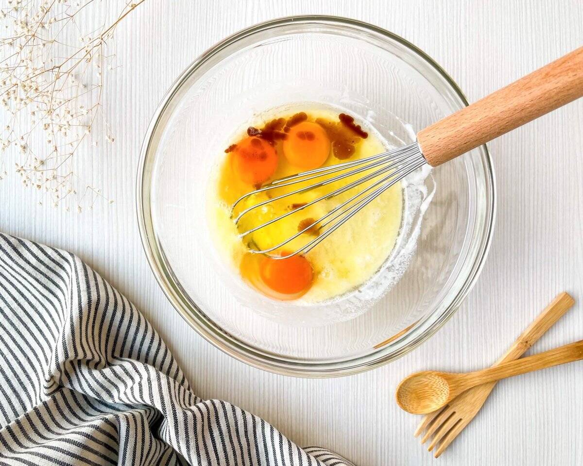 A glass bowl containing eggs and a whisk is placed on a light surface, accompanied by wooden utensils and a striped cloth, suggesting preparation for a Dutch baby pancake recipe or other breakfast brunch ideas.
