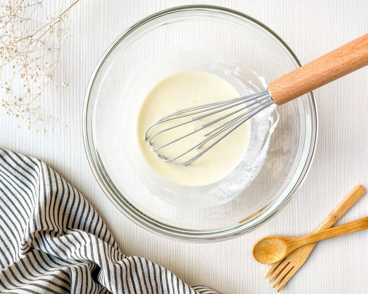 A glass bowl containing a batter mixture, with a whisk resting inside, accompanied by a striped kitchen towel and wooden utensils, set against a light wooden surface, indicating preparation for a Dutch baby pancake recipe.
