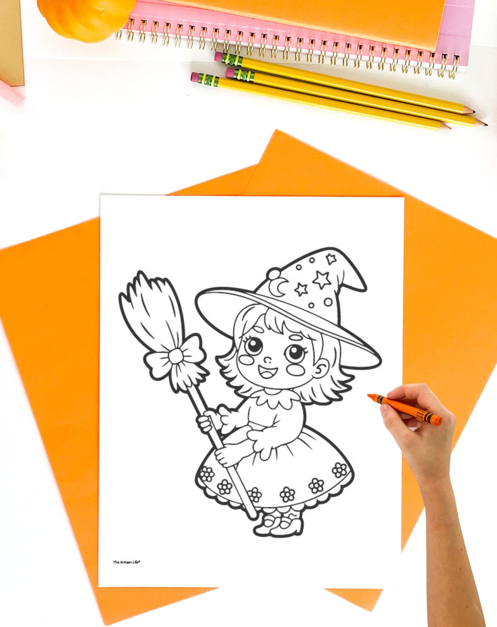 A hand coloring a cute witch illustration on a black and white Halloween coloring page, surrounded by colorful papers and stationery.
