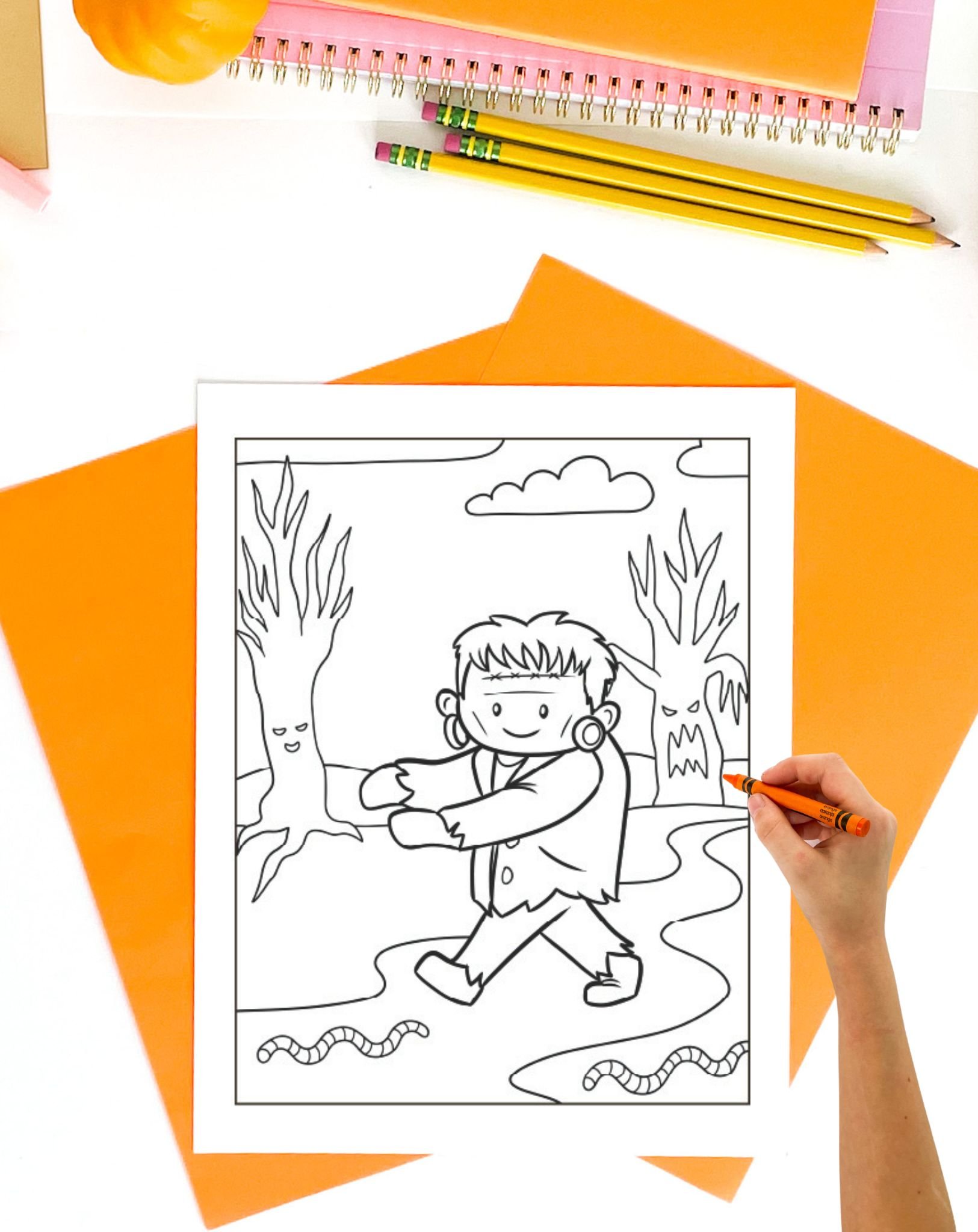 A child is coloring a printable Halloween page featuring a cartoon-style Frankenstein character walking among trees with a ghostly figure in the background.

