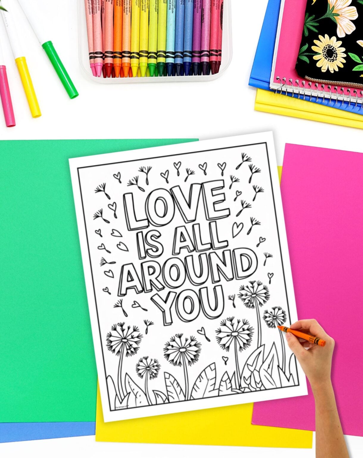 A hand is coloring a printable page featuring the phrase "LOVE IS ALL AROUND YOU," surrounded by dandelions and hearts, on a workspace with various colorful sheets and art supplies, highlighting mindfulness and kindness activities for kids.