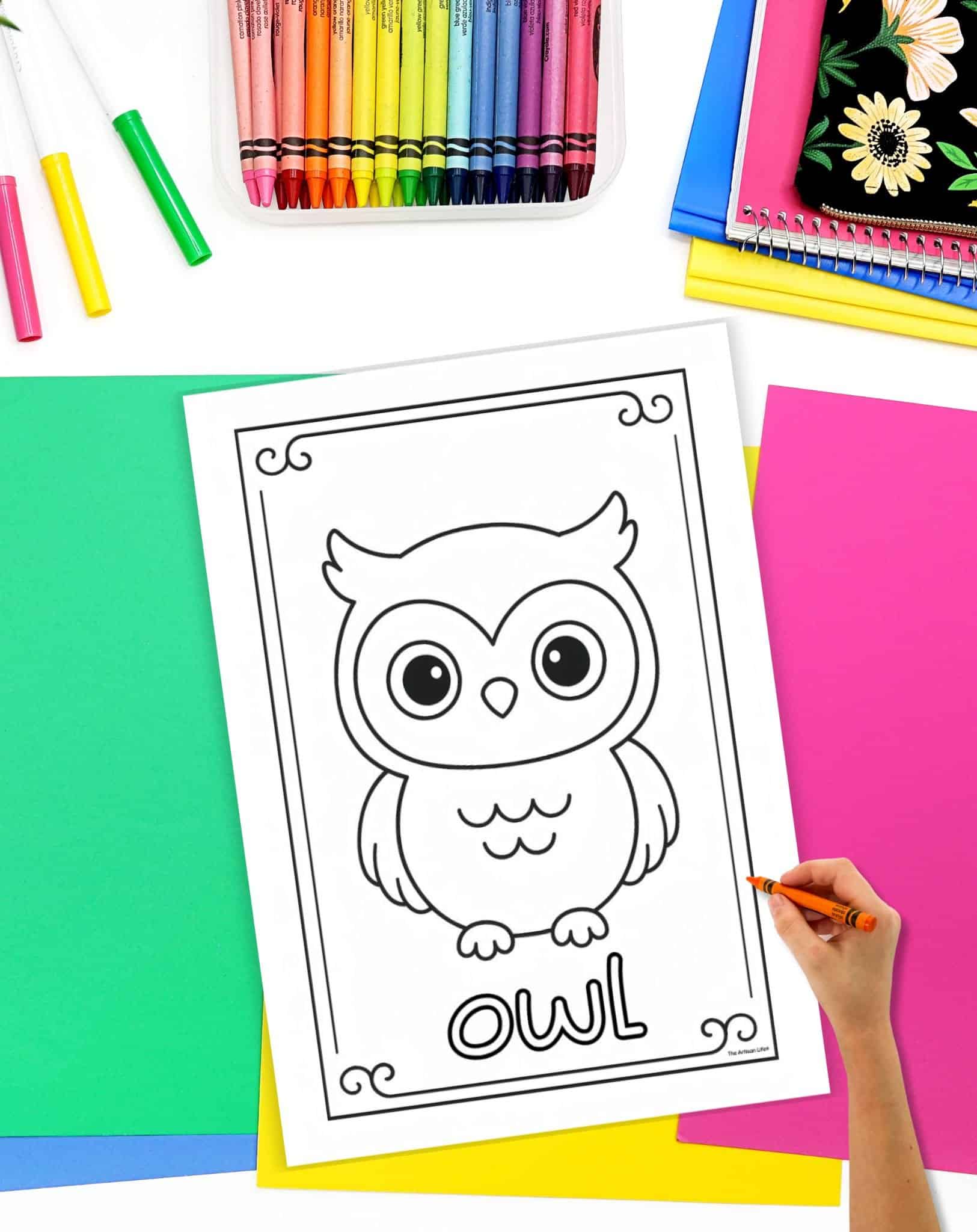 A hand is holding an orange crayon as it colors a black-and-white printable page featuring a cartoon owl, surrounded by various colored papers and a container of crayons.
