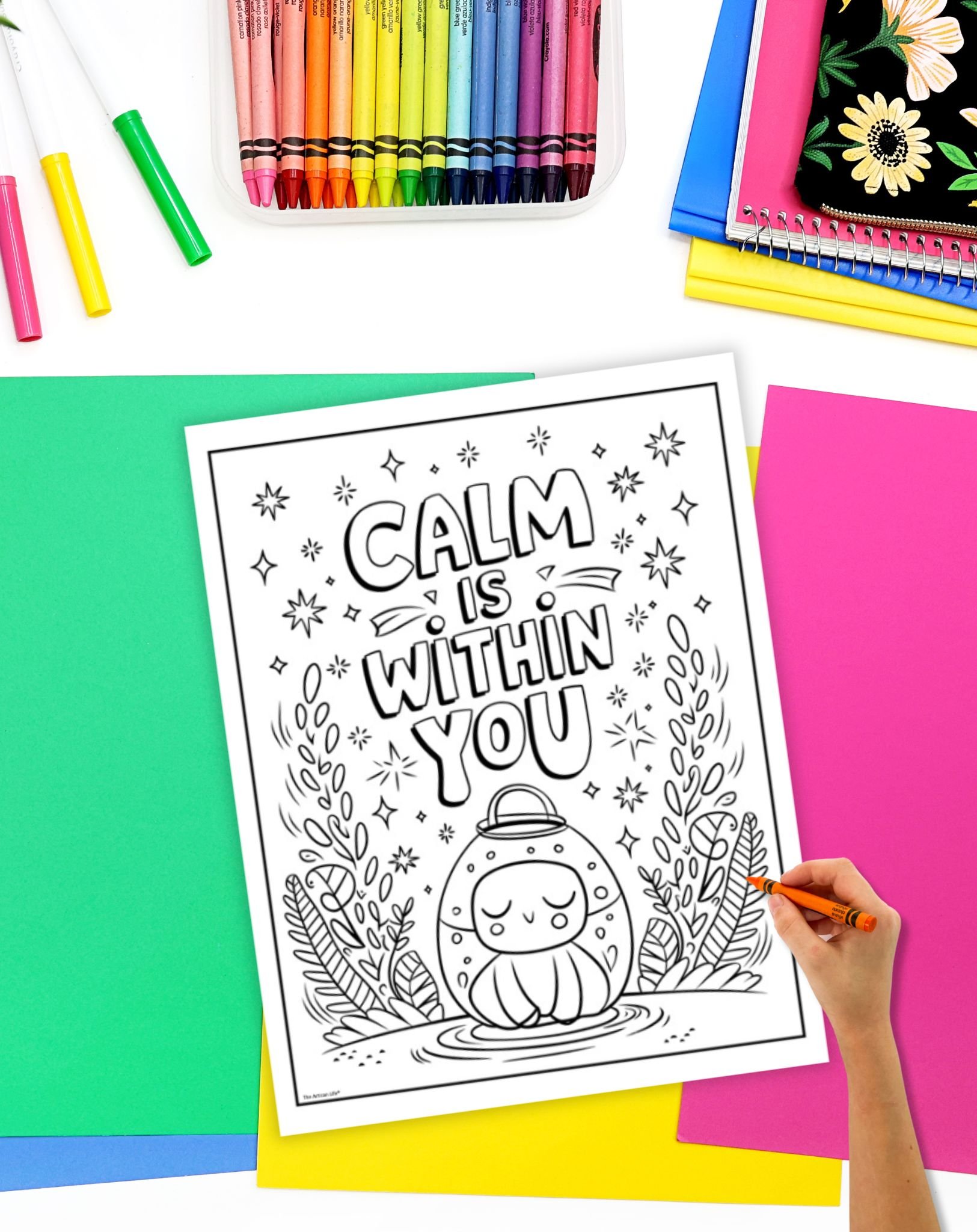 A hand colors in a printable mindfulness coloring page featuring the phrase "Calm is Within You," surrounded by a whimsical design, against a backdrop of colorful art supplies and sheets of paper.
