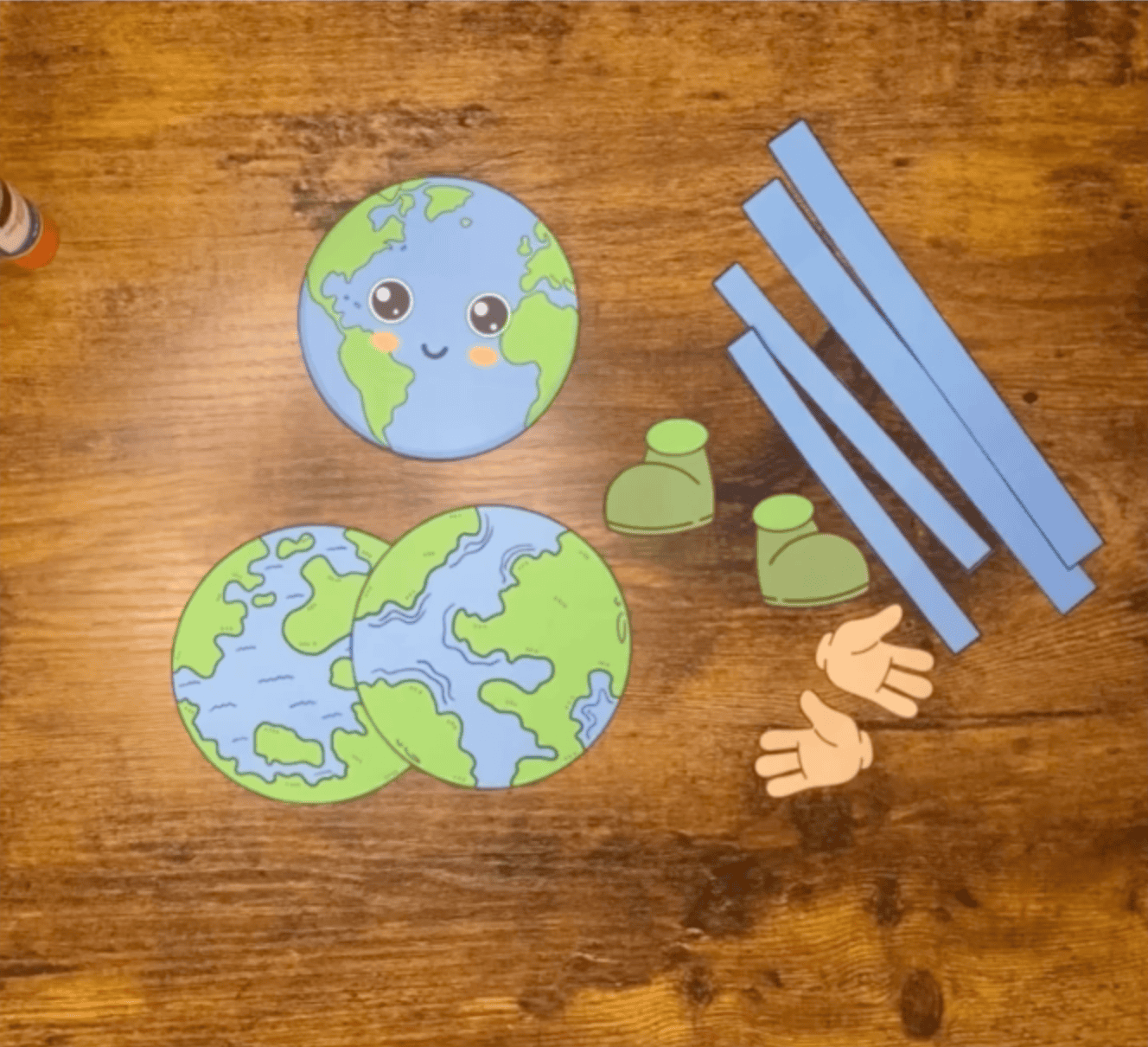 A paper craft of a smiling Earth character with flexible arms and legs, designed as a simple Earth Day craft for preschool and elementary school children, is displayed on a wooden table alongside a glue stick.
