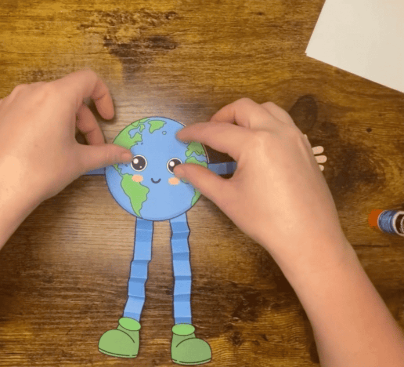 A person is assembling a cheerful Earth character with paper arms and legs as part of simple Earth Day crafts aimed at preschoolers and young children.
