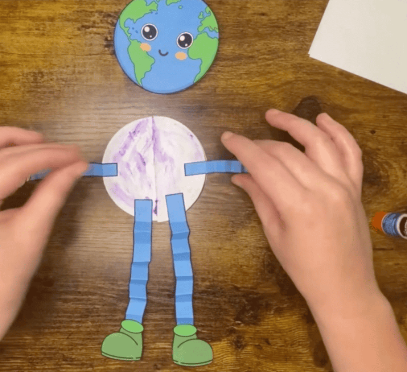 A pair of hands assembles a colorful Earth-themed craft featuring a circular Earth shape on top of a character body made of paper, representing simple Earth Day crafts for kids.
