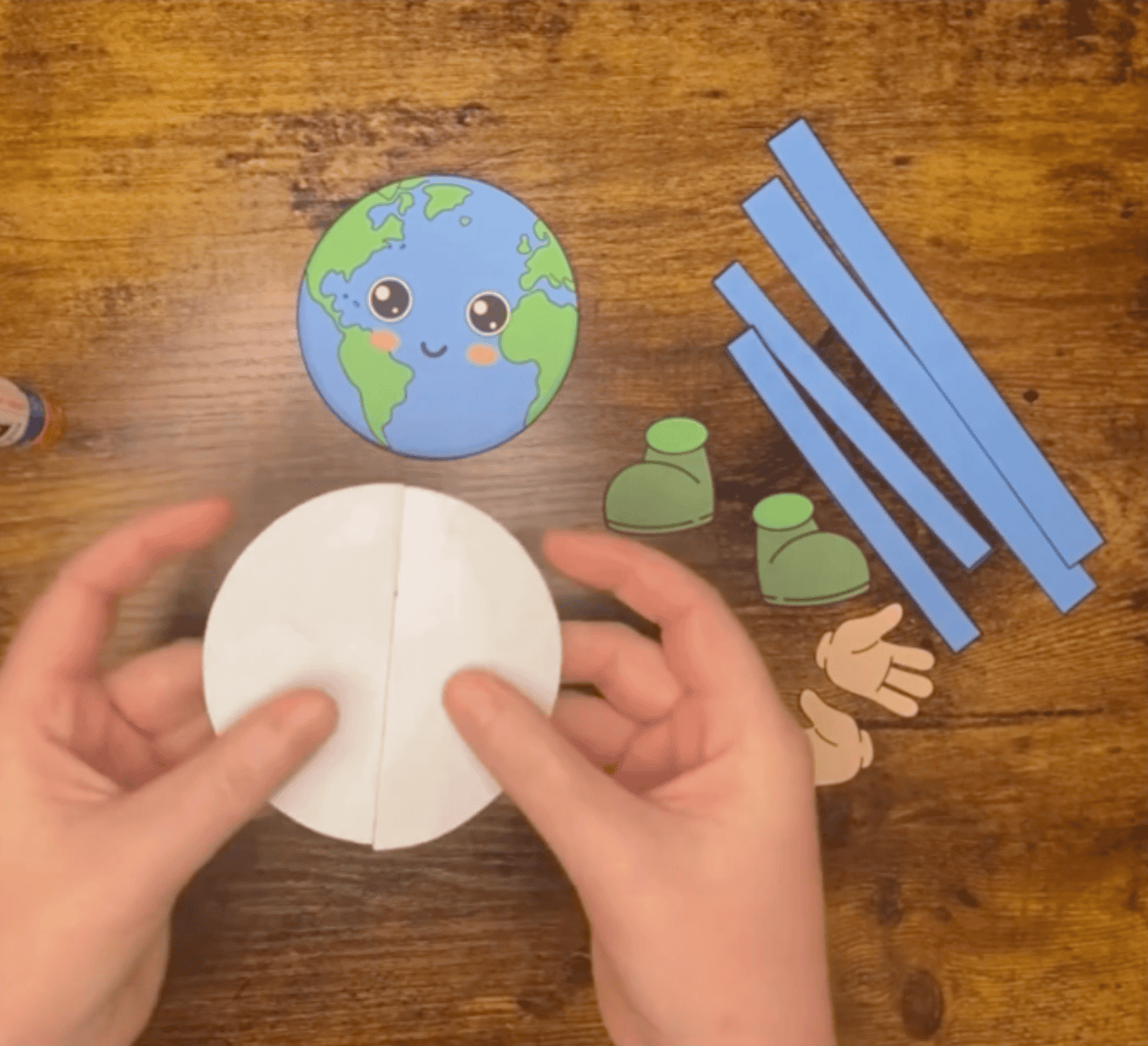 A pair of hands holds a folded white circle, with a colorful Earth template and various craft supplies, including green boots and blue strips, set on a wooden surface, illustrating simple Earth Day crafts for kids.
