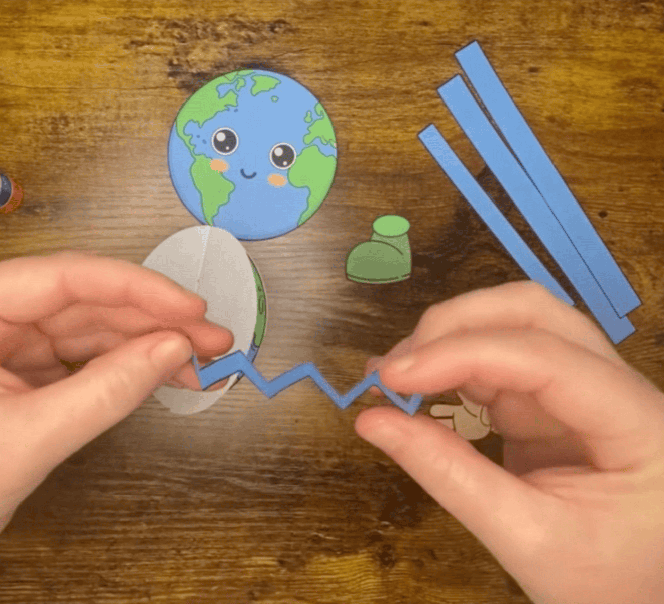 A person's hands are shown assembling simple Earth Day crafts for kids, featuring a colorful paper Earth, strips of blue paper, and a green boot, suitable for preschool and elementary school activities.
