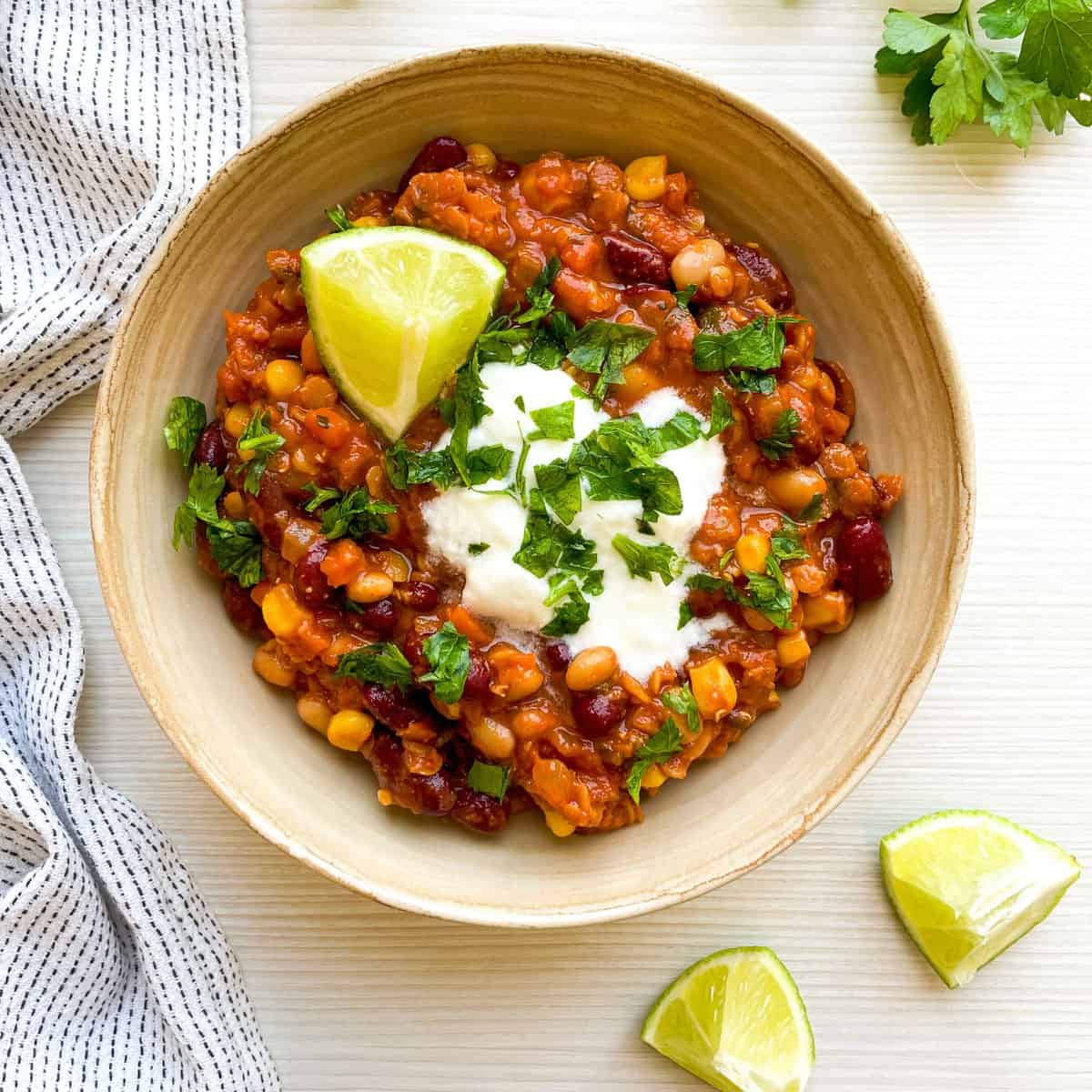 A bowl of three bean chili topped with cilantro and sour cream, accompanied by lime wedges, representing an easy vegetarian chili recipe that is nutritious and filling.
