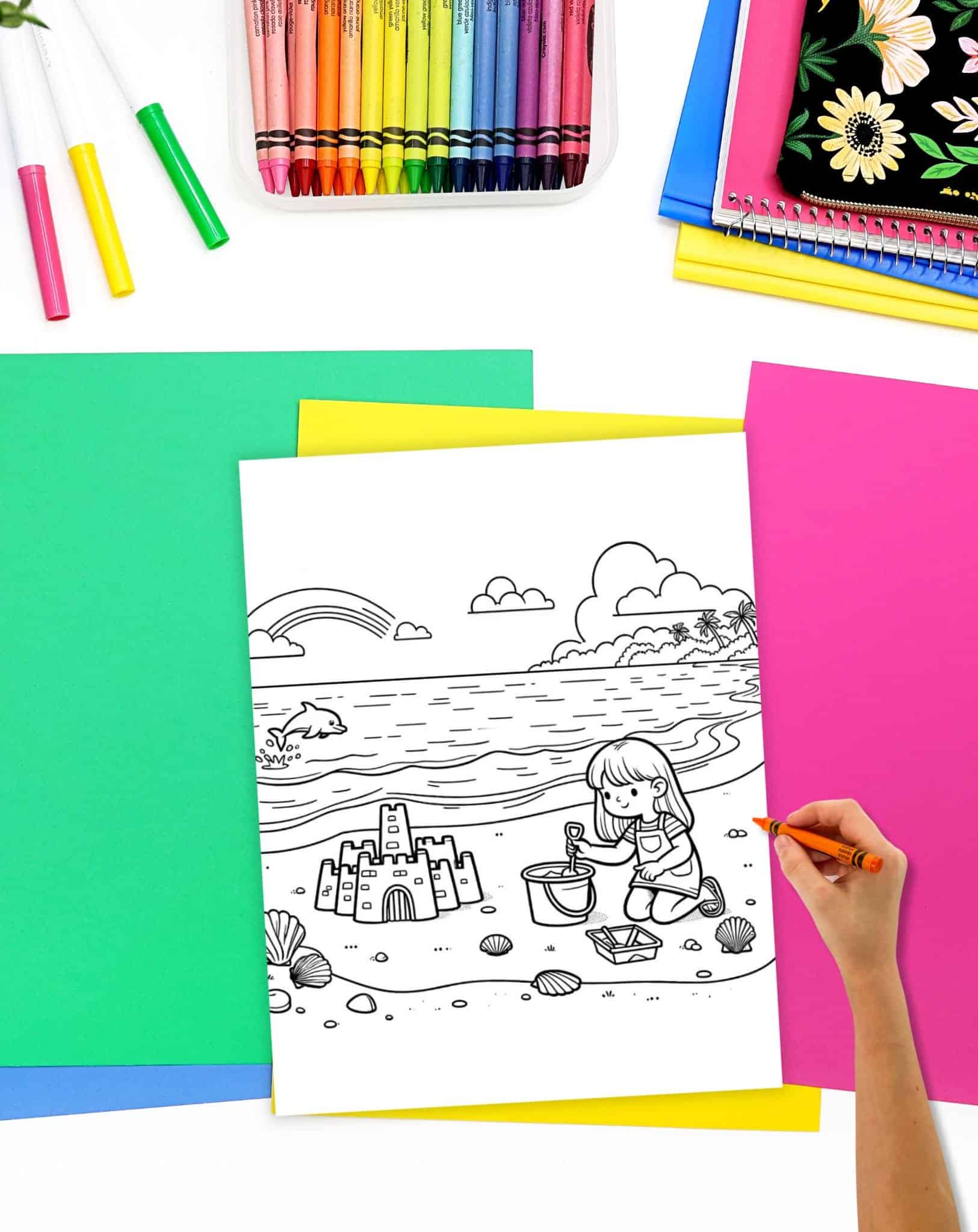 A child is coloring a beach scene featuring a sandcastle on a white coloring page, surrounded by colorful art supplies and sheets of construction paper, highlighting summer-themed activities.
