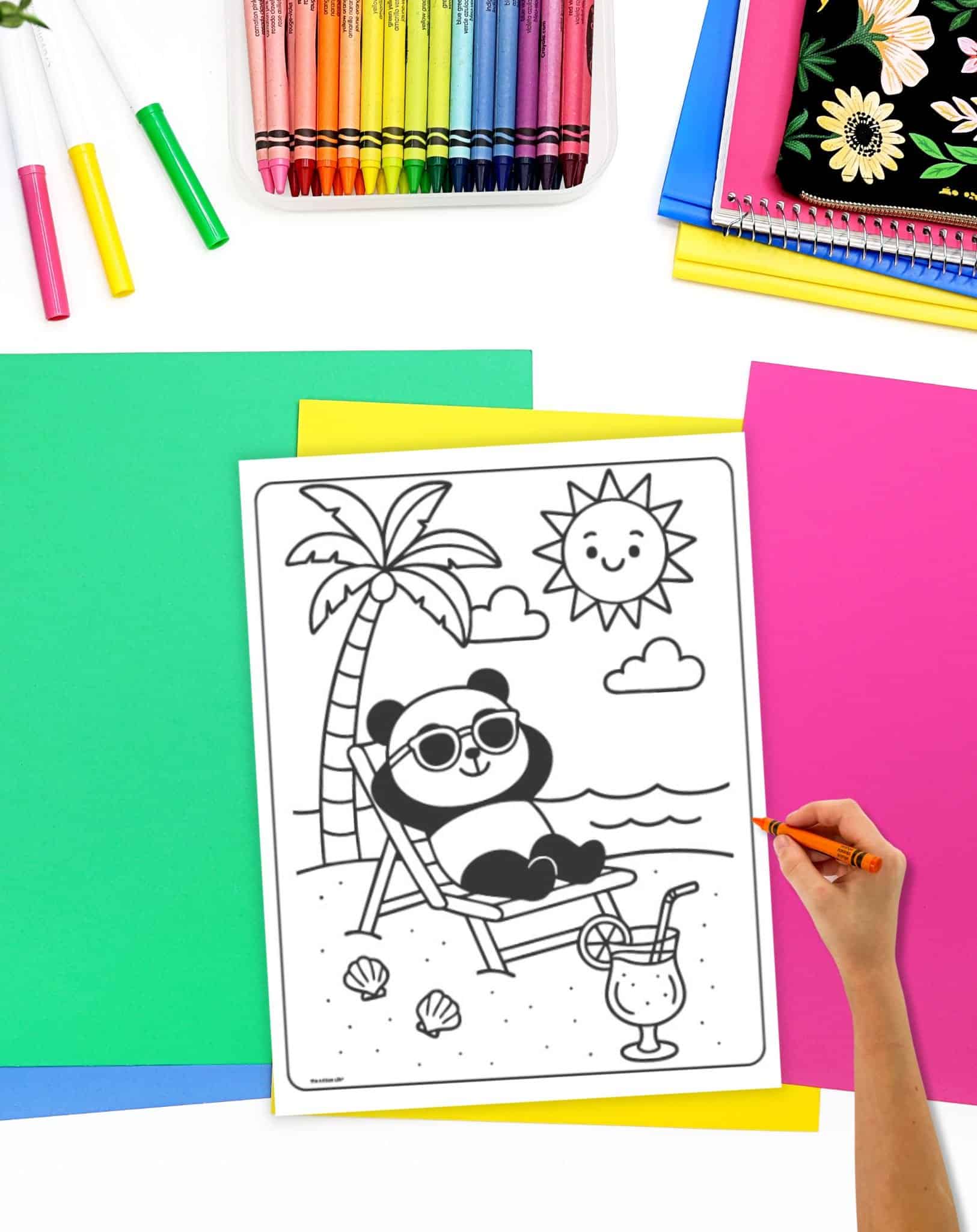 A hand holding an orange crayon is coloring a bold and easy summer coloring page featuring a panda wearing sunglasses relaxing on a beach chair with a drink, surrounded by a palm tree and a sun, with various colorful crayons and paper in the background.
