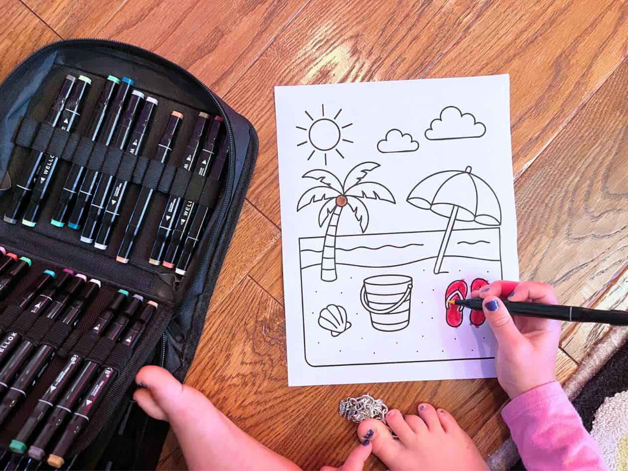 A child is coloring a beach-themed coloring page featuring palm trees, a sun, and flip-flops, using markers from a nearby case on a wooden floor.
