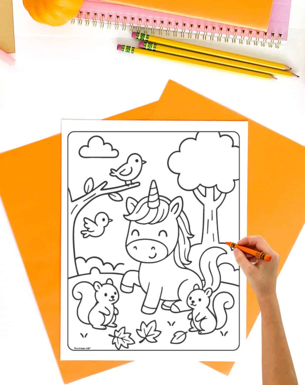 A person is coloring a free printable fall coloring page featuring a unicorn, two squirrels, birds, and autumn leaves on a white sheet surrounded by orange stationery.