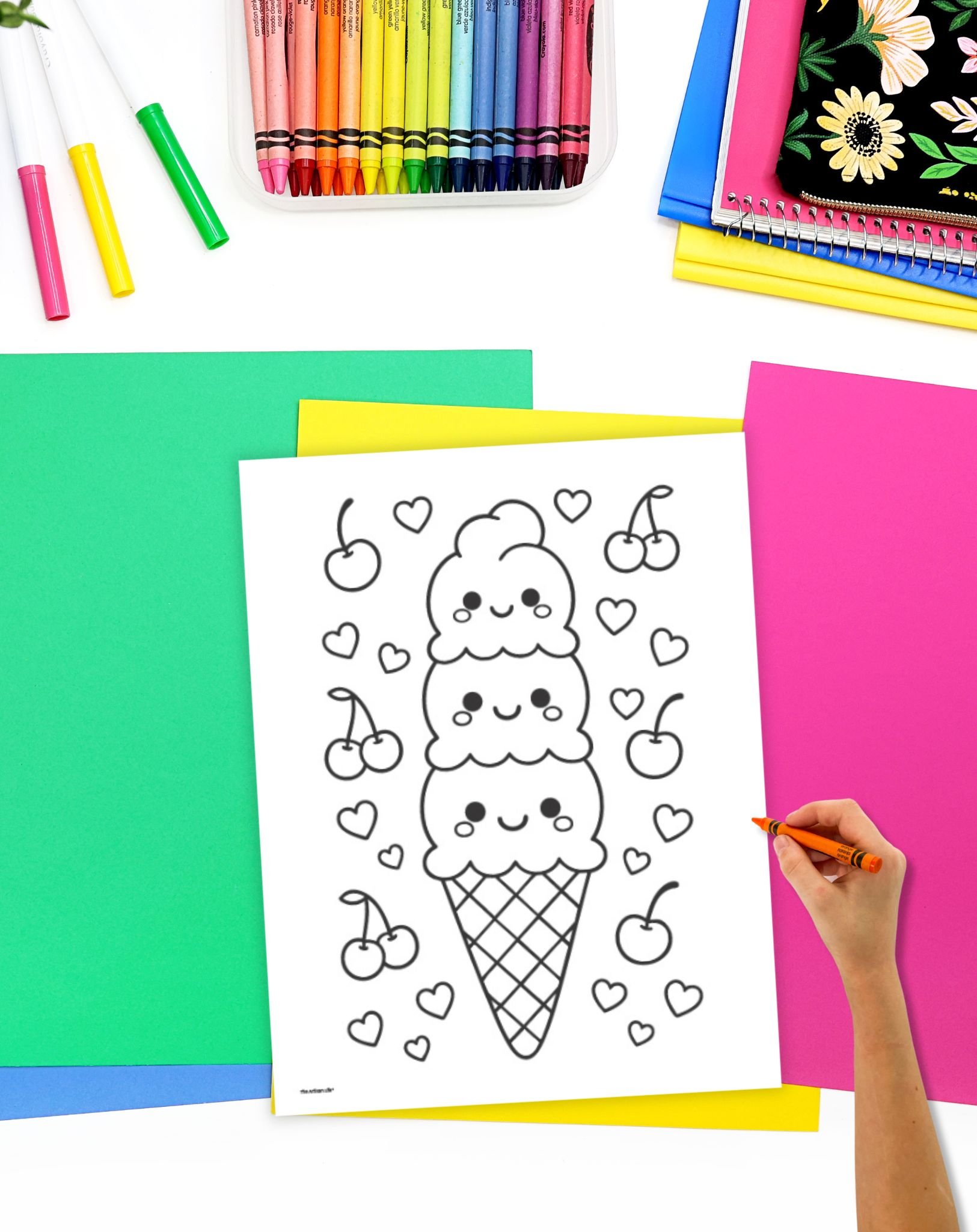A hand is coloring a black-and-white ice cream sundae coloring page, surrounded by colorful construction paper and a box of crayons, representing fun summer activities for kids.
