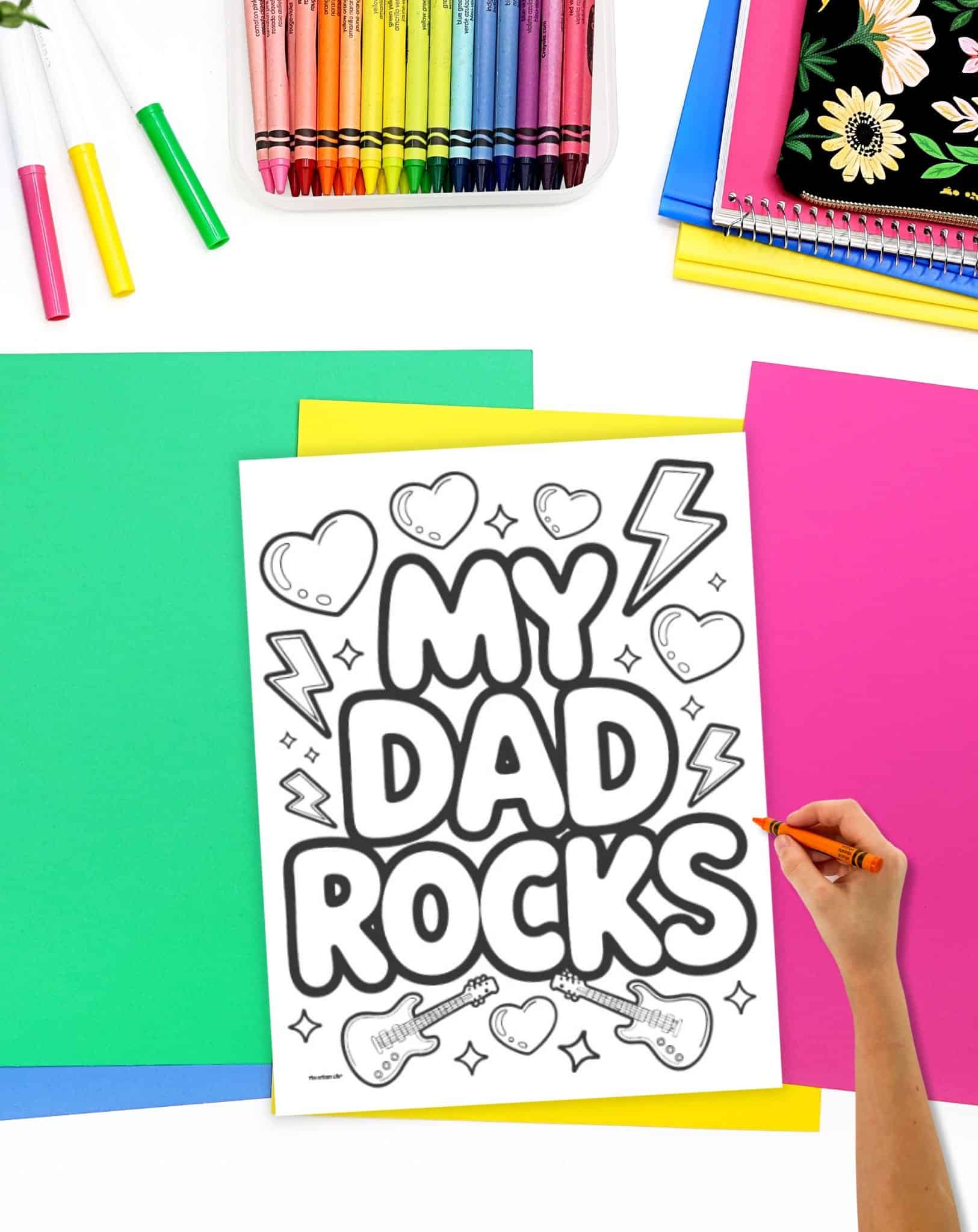 A hand is coloring a black and white Father's Day coloring page that reads "MY DAD ROCKS," surrounded by crayons and colorful paper, highlighting it as a part of free printable Father’s Day coloring pages and easy Father’s Day crafts.
