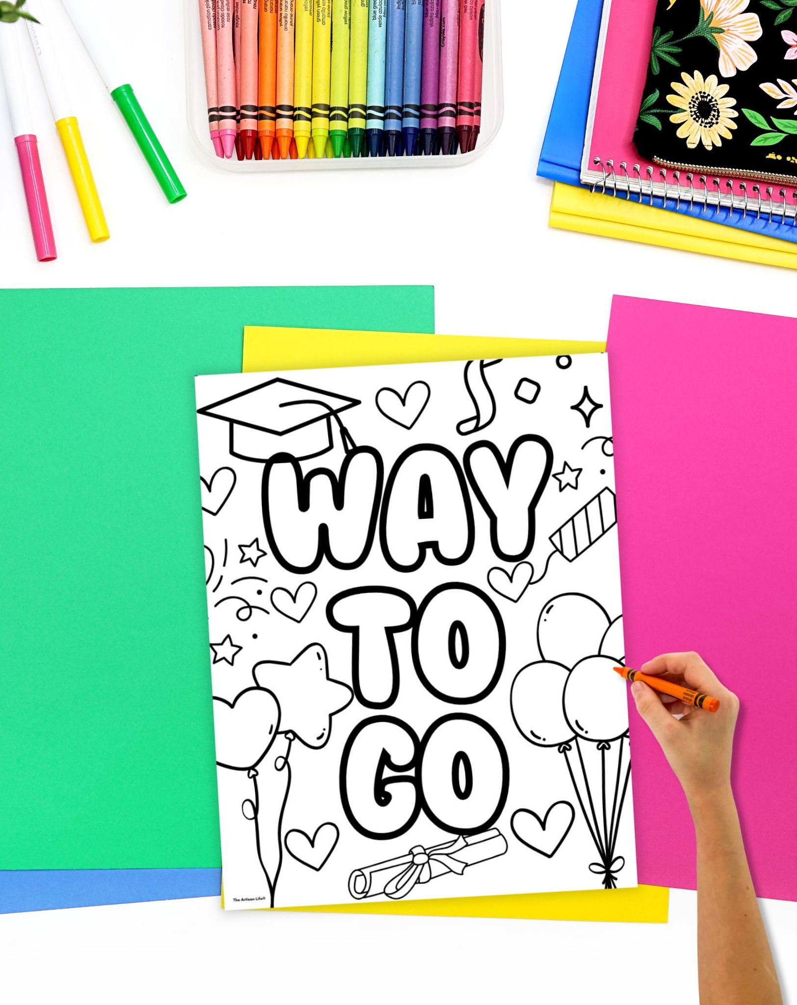 A hand is coloring a "WAY TO GO" graduation-themed coloring page surrounded by colorful crayons and various sheets of paper, suitable for preschool graduation activities and crafts.
