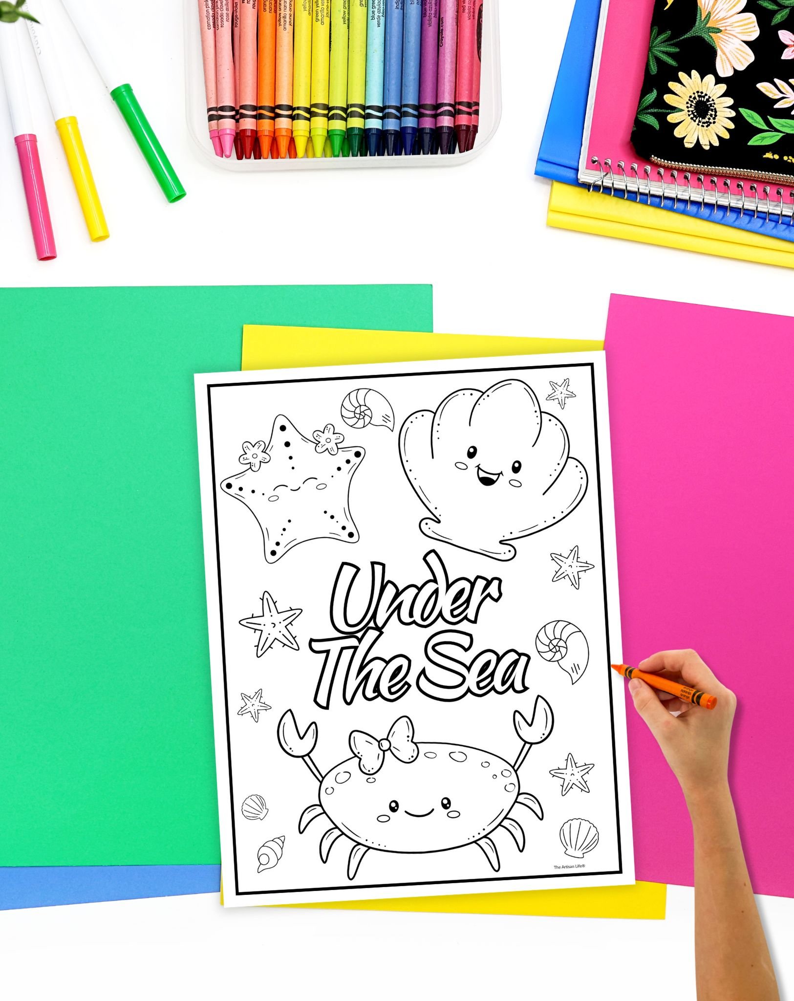 A hand is coloring a printable summer coloring page featuring marine life, including a crab and starfish, surrounded by colored crayons and construction paper.
