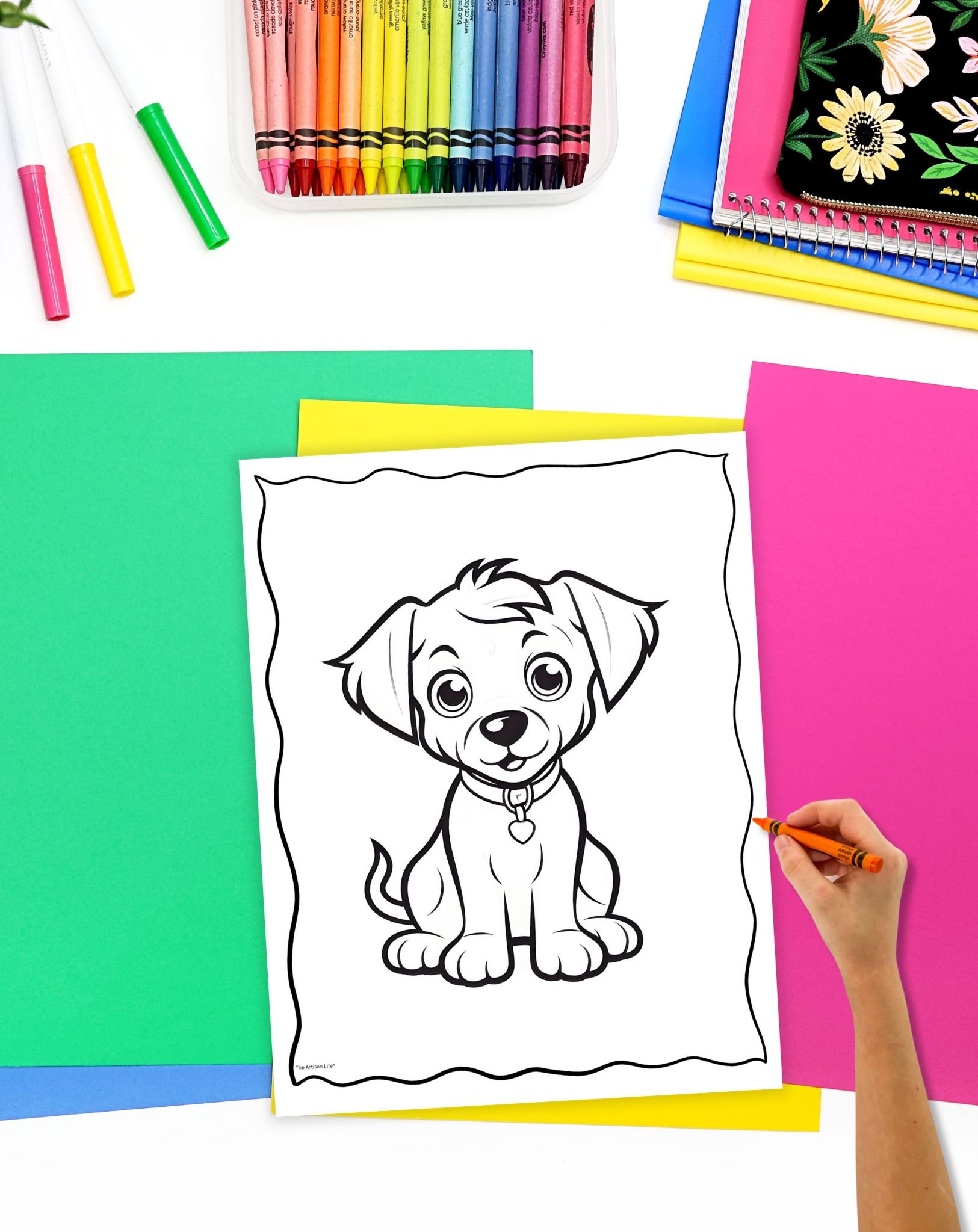 A hand is coloring in a printable dog coloring sheet featuring a cartoon puppy, surrounded by colorful papers and crayons, ideal for kids' art activities.
