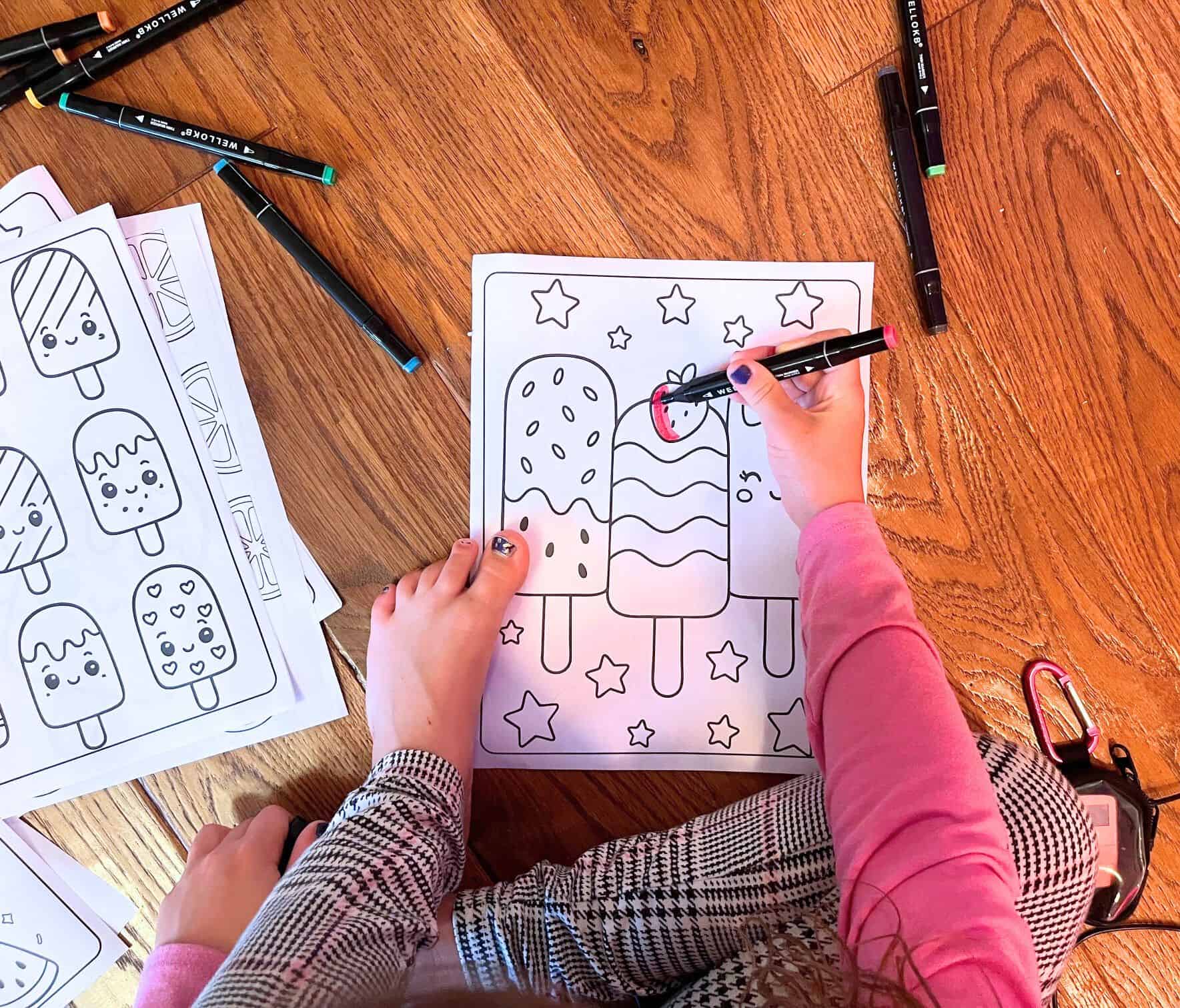 A child colors a cute popsicle-themed page with stars, surrounded by summer-themed coloring pages and markers on a wooden floor.

