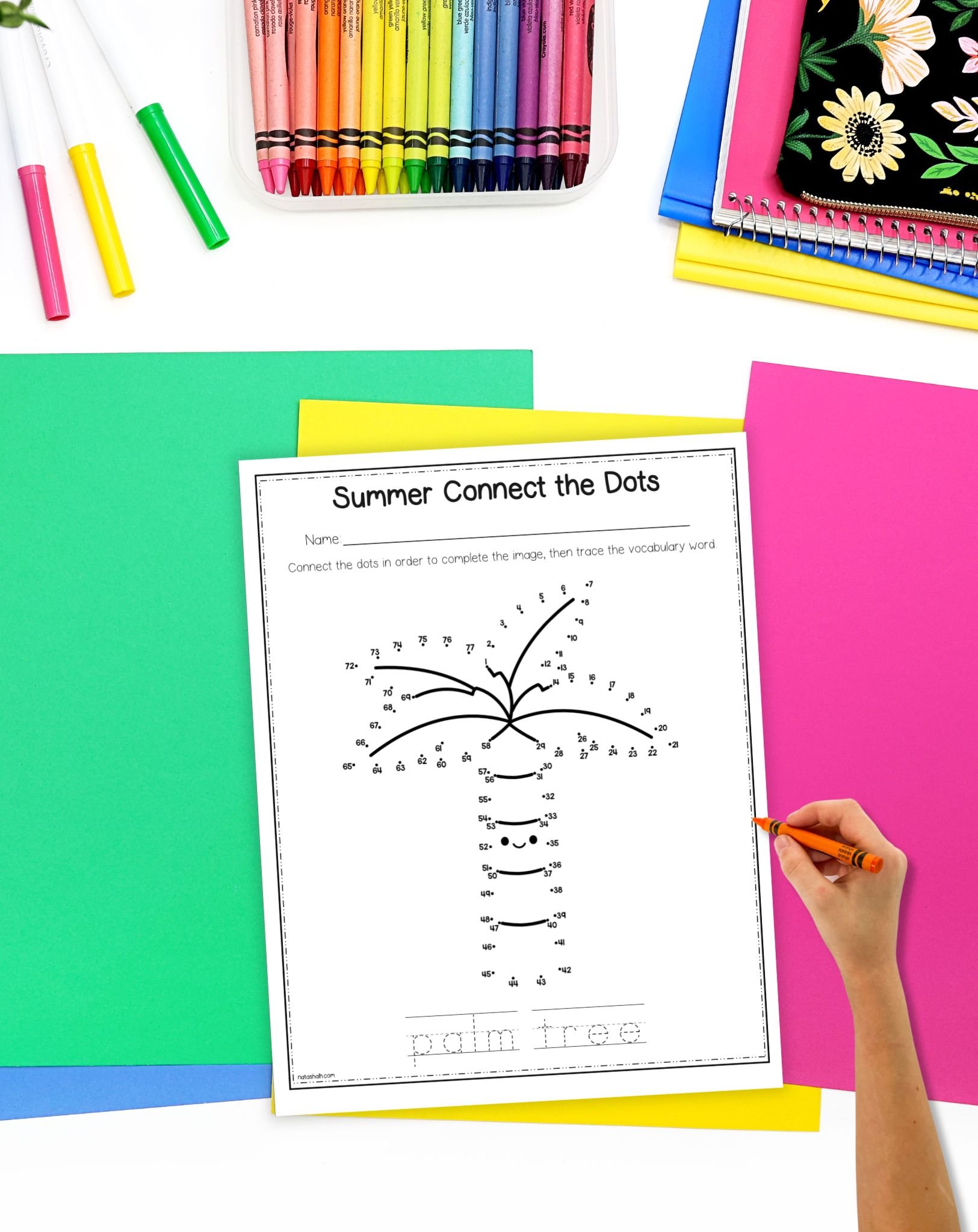 A hand is completing a "Summer Connect the Dots" worksheet featuring a palm tree, surrounded by colorful papers and crayons, suitable for kids' summer activities and math worksheets.
