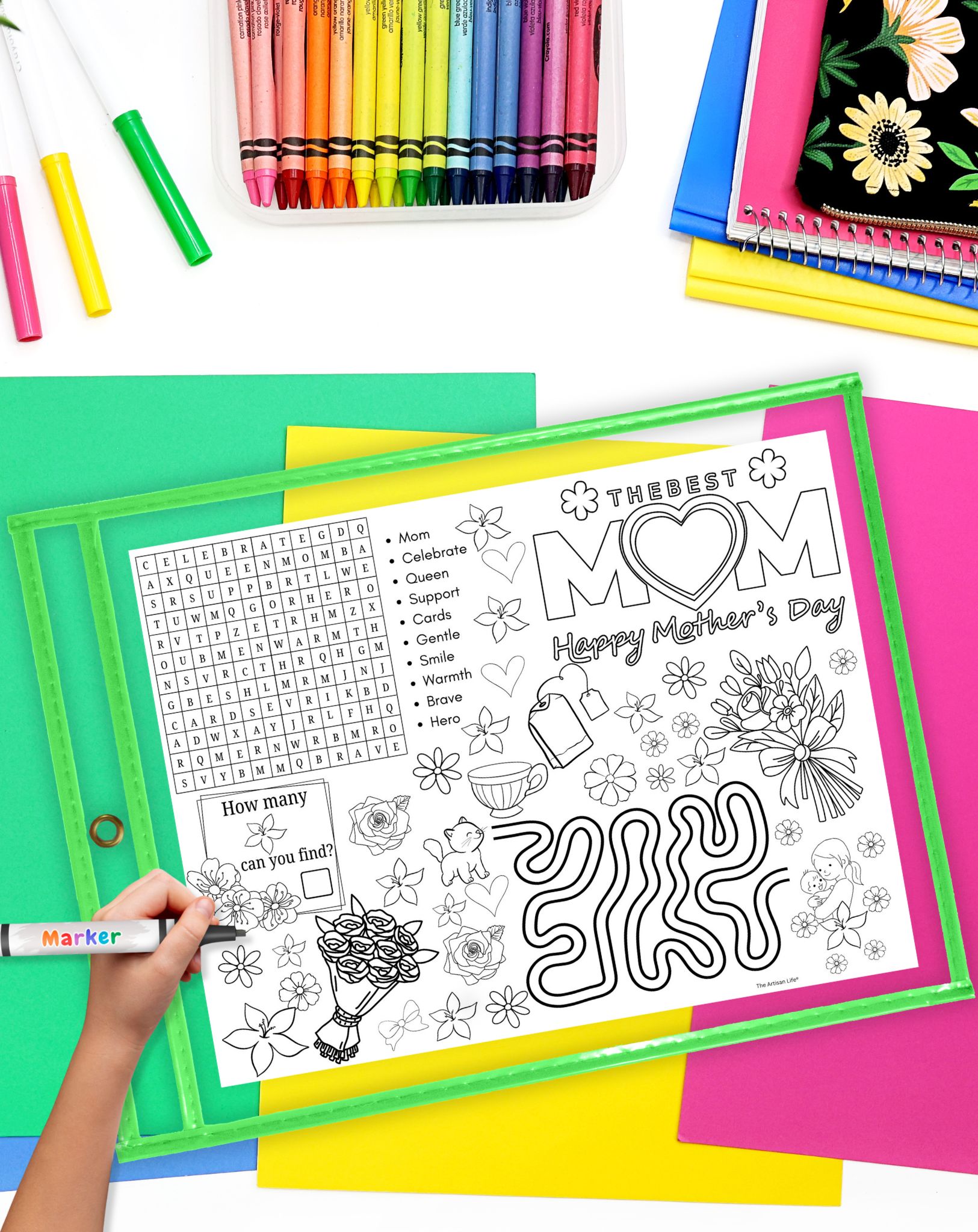A child's hand colors a Mother's Day printable activity mat featuring word searches, coloring images, and prompts for fun printable activities, surrounded by colorful crayons and sheets of paper.
