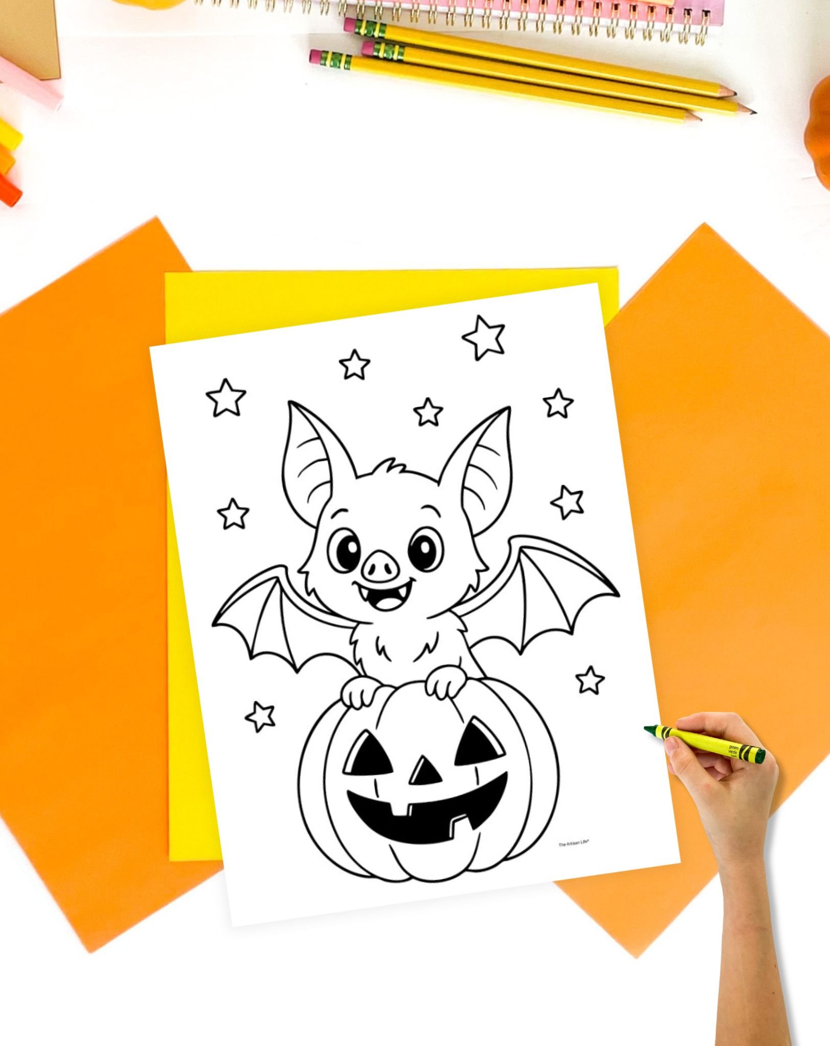 A person's hand holds a crayon above a printable coloring page featuring a cute bat sitting on a jack-o'-lantern, surrounded by stars, with colors and craft supplies in the background, ideal for bat coloring sheets or free Halloween coloring pages.
