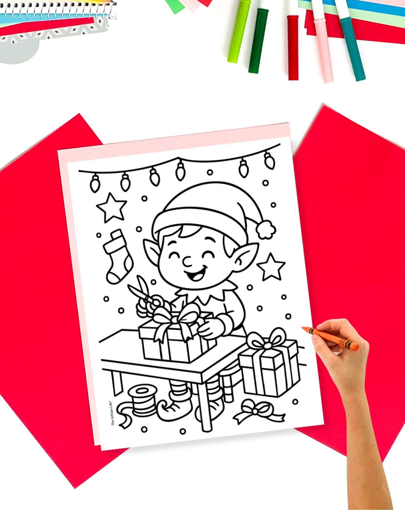 A hand holds a crayon near a black-and-white coloring page depicting a cheerful elf in Santa's workshop, surrounded by gifts, in a scene ideal for free printable Christmas coloring pages.
