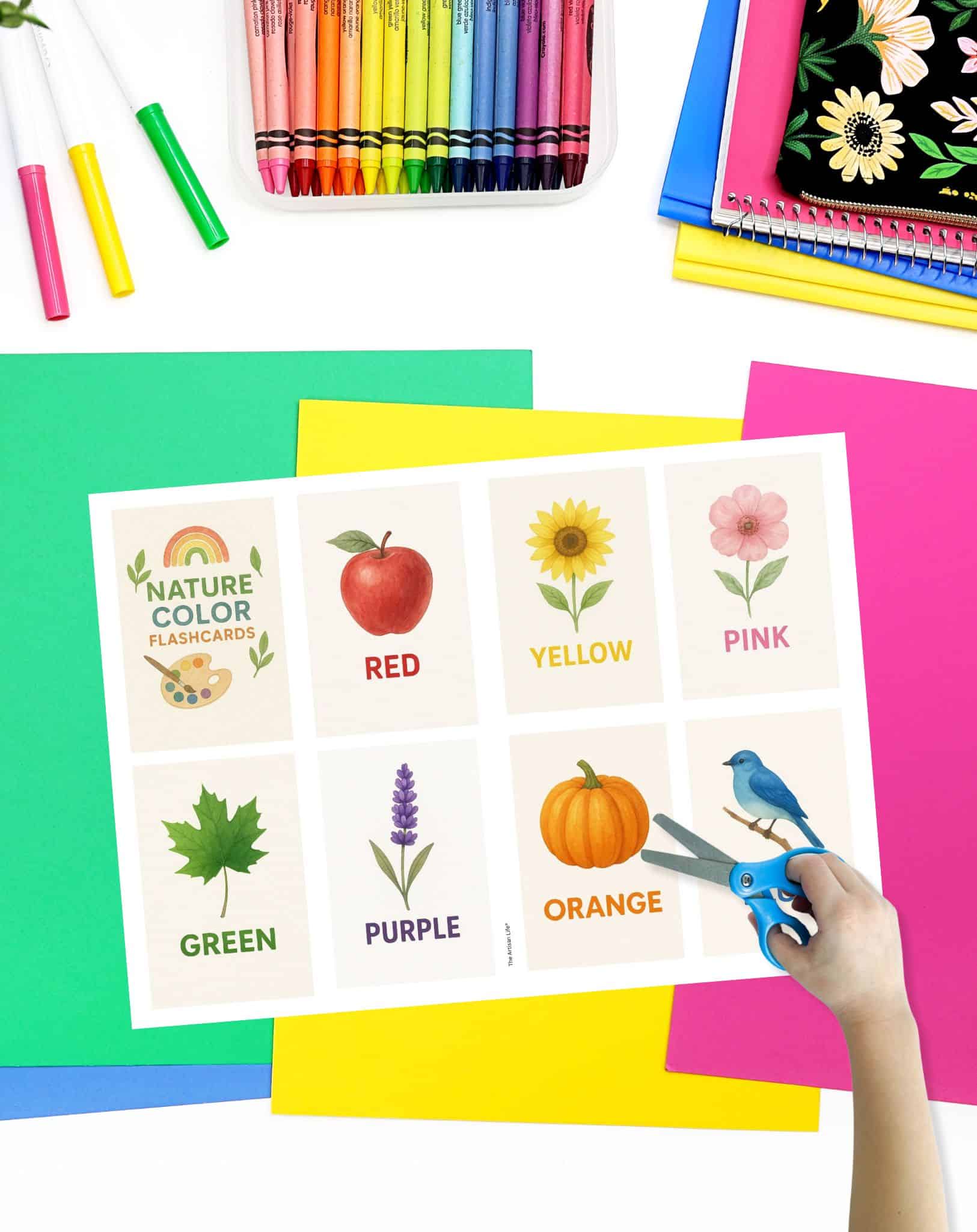 A hand holds scissors near a sheet of color flashcards featuring illustrations of various colors and objects, surrounded by colorful cardstock and markers, suitable for preschoolers and toddlers.

