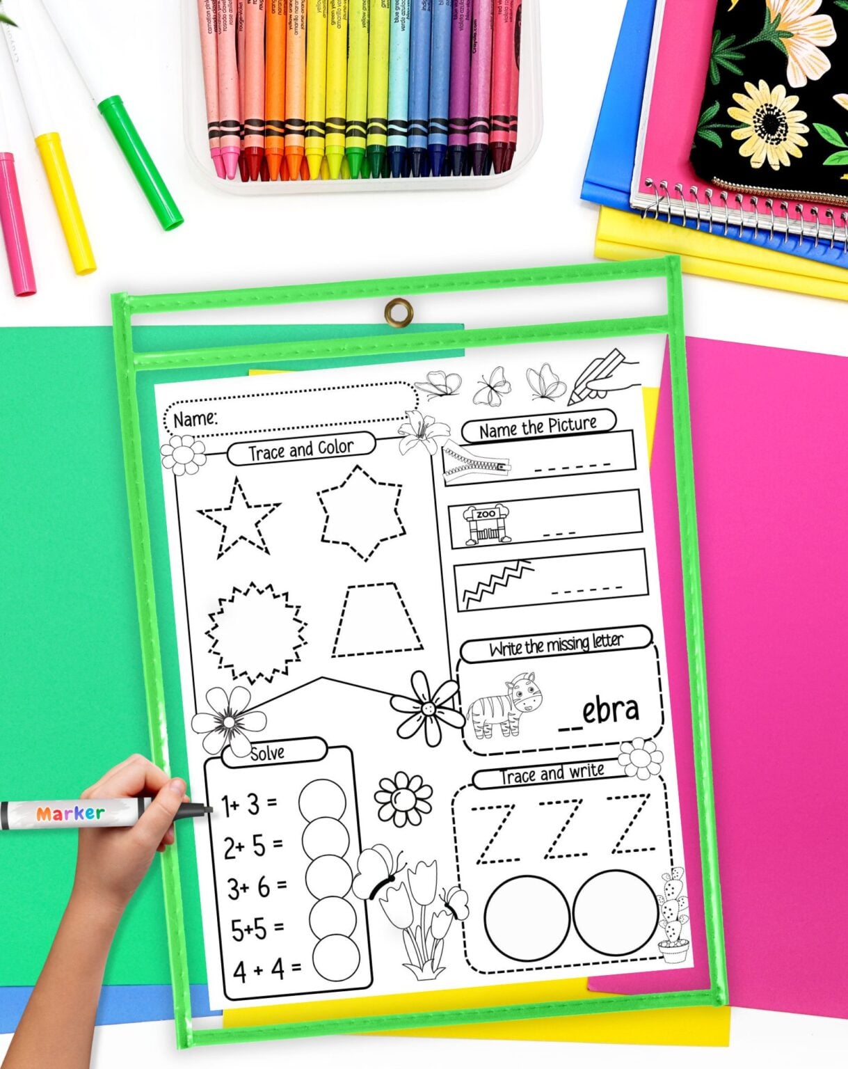 A child's hand is holding a marker and completing a kindergarten worksheet focused on math and writing skills, surrounded by colorful crayons and sheets of paper, suitable for morning work in a homeschool or classroom setting.