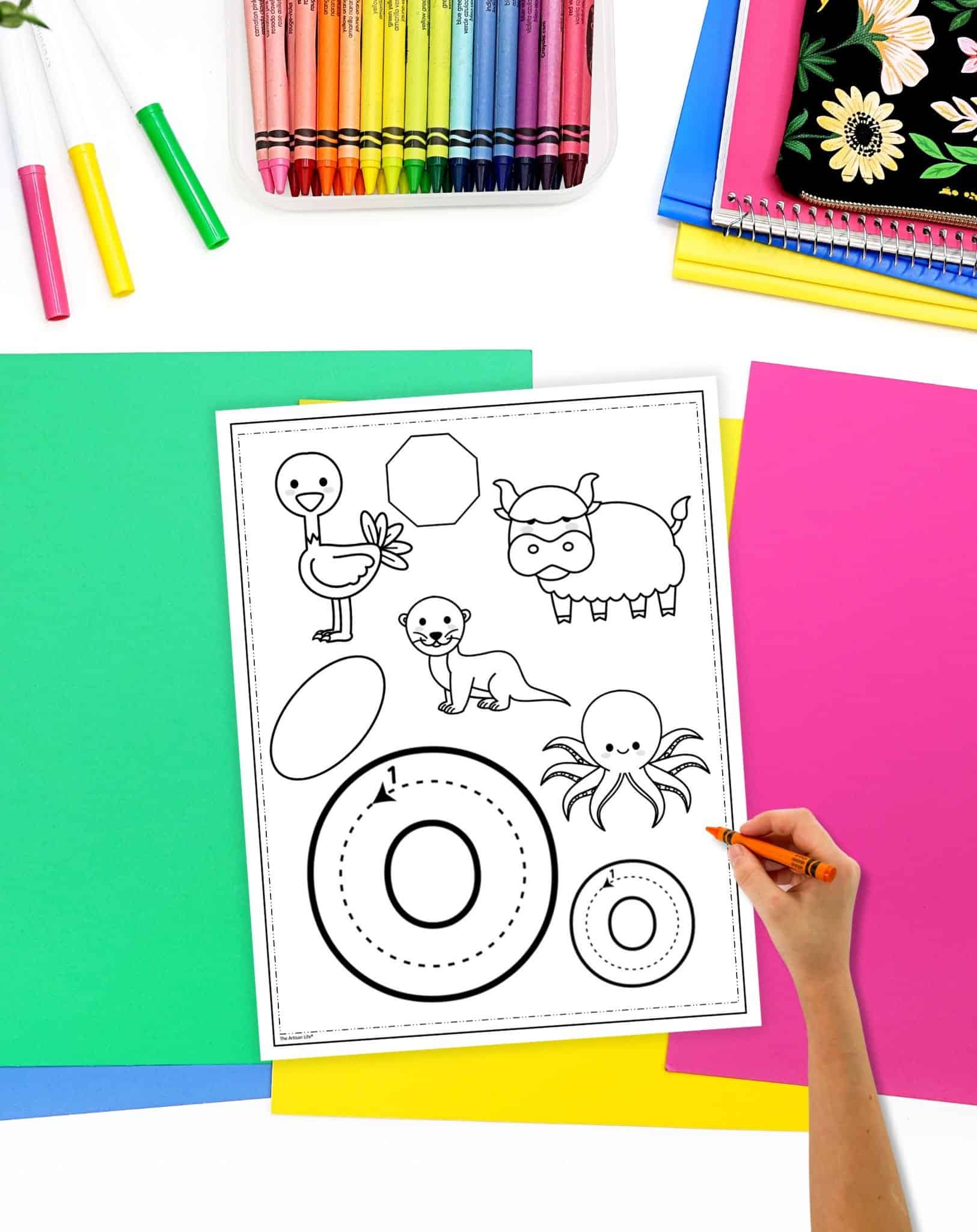 A hand is holding an orange crayon above a black and white alphabet coloring page featuring various animals and shapes, surrounded by colorful sheets of paper and a set of crayons.
