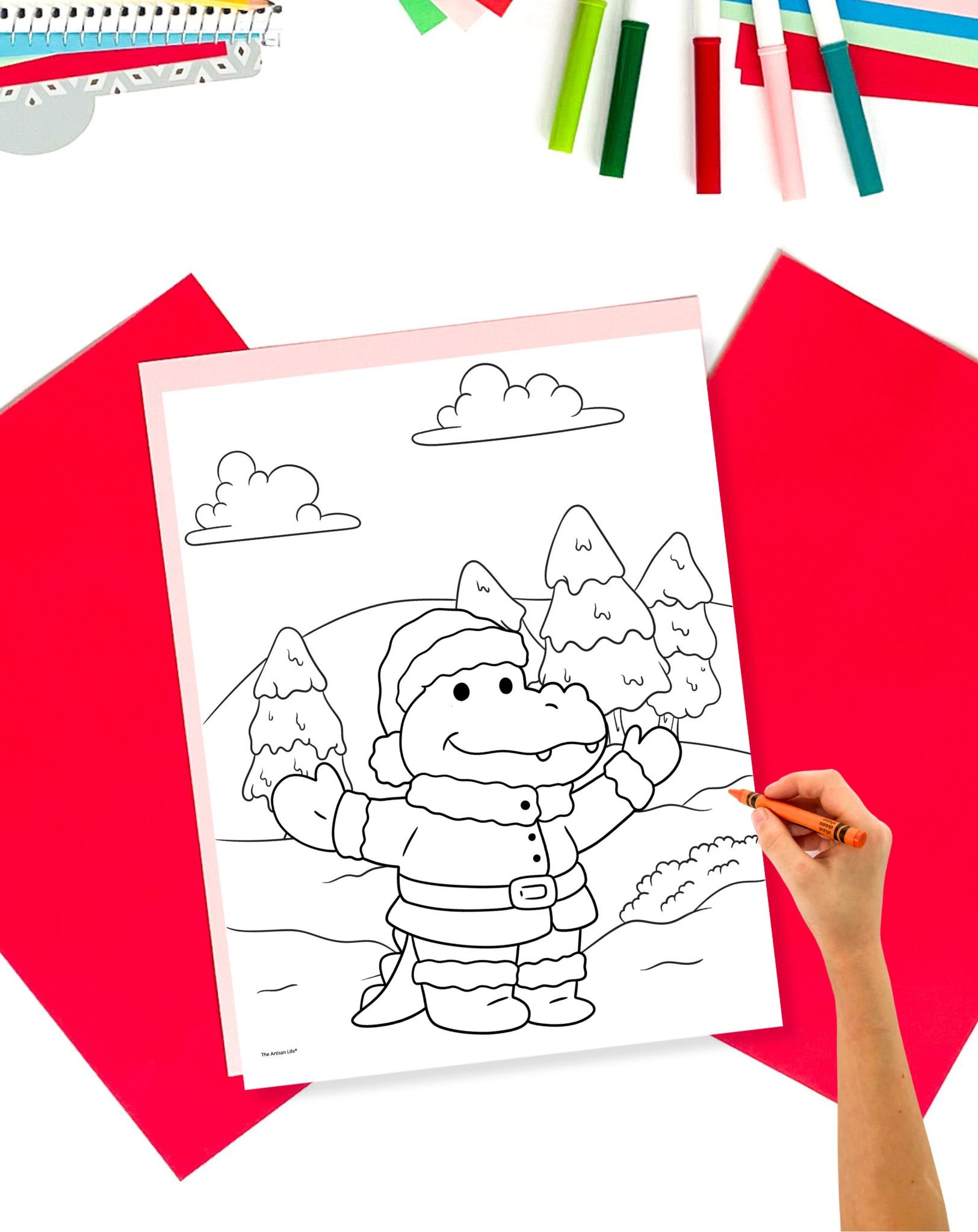 A hand colors a bold Christmas coloring page featuring a cartoon alligator in a Santa outfit, surrounded by trees and hills, with additional coloring materials and paper in the background.
