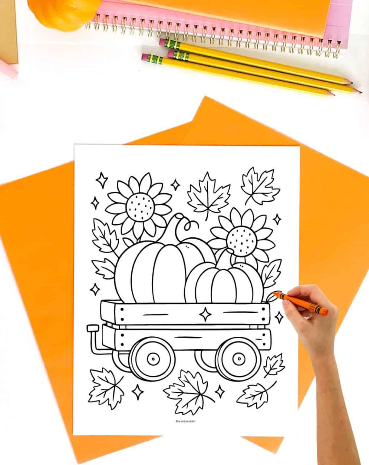 A hand holds a crayon while coloring a printable fall-themed coloring page depicting a wagon filled with pumpkins, sunflowers, and autumn leaves, surrounded by orange sheets of paper and pencils.