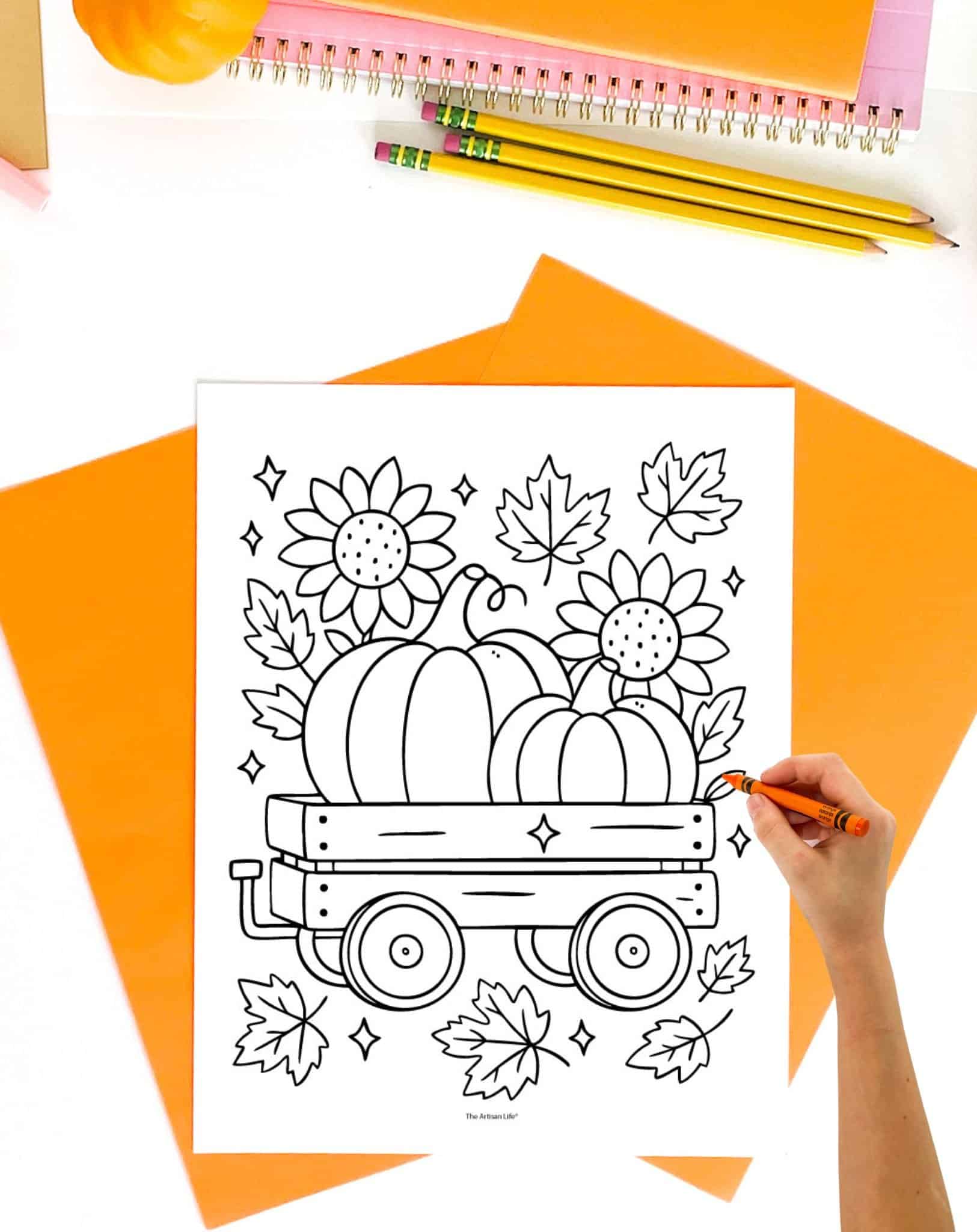 A hand holds a crayon while coloring a printable fall-themed coloring page depicting a wagon filled with pumpkins, sunflowers, and autumn leaves, surrounded by orange sheets of paper and pencils.

