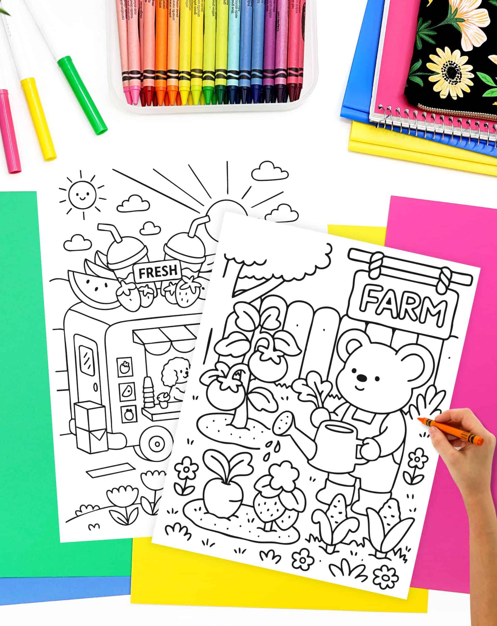 An assortment of colorful crayons is displayed alongside two printable coloring pages featuring a summer theme, one depicting a fruit stand and the other a bear watering a garden, set against a backdrop of vibrant construction paper.
