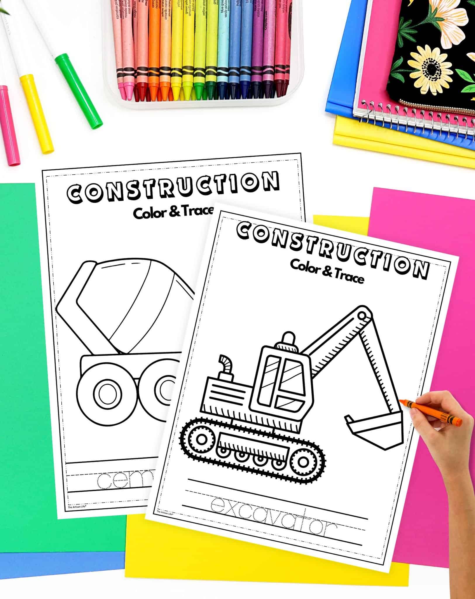 A child’s hand colors a printable construction-themed coloring page featuring a dump truck and an excavator, surrounded by various colored crayons and sheets of construction paper.
